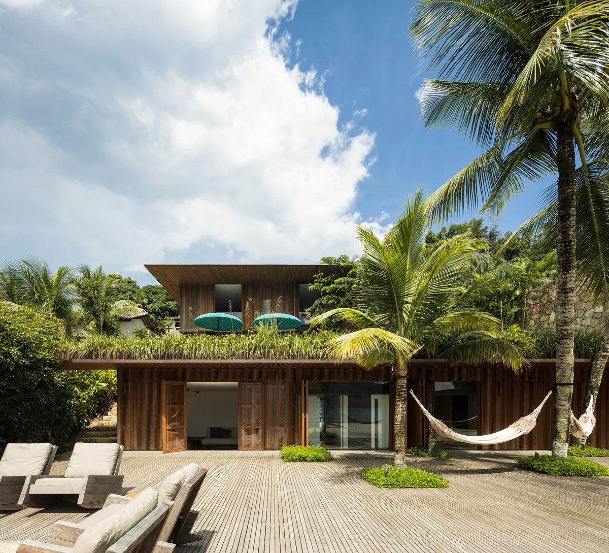 A modern tropical home features a slatted wood exterior, a spacious wooden deck with integrated palm trees, and lounging hammocks.