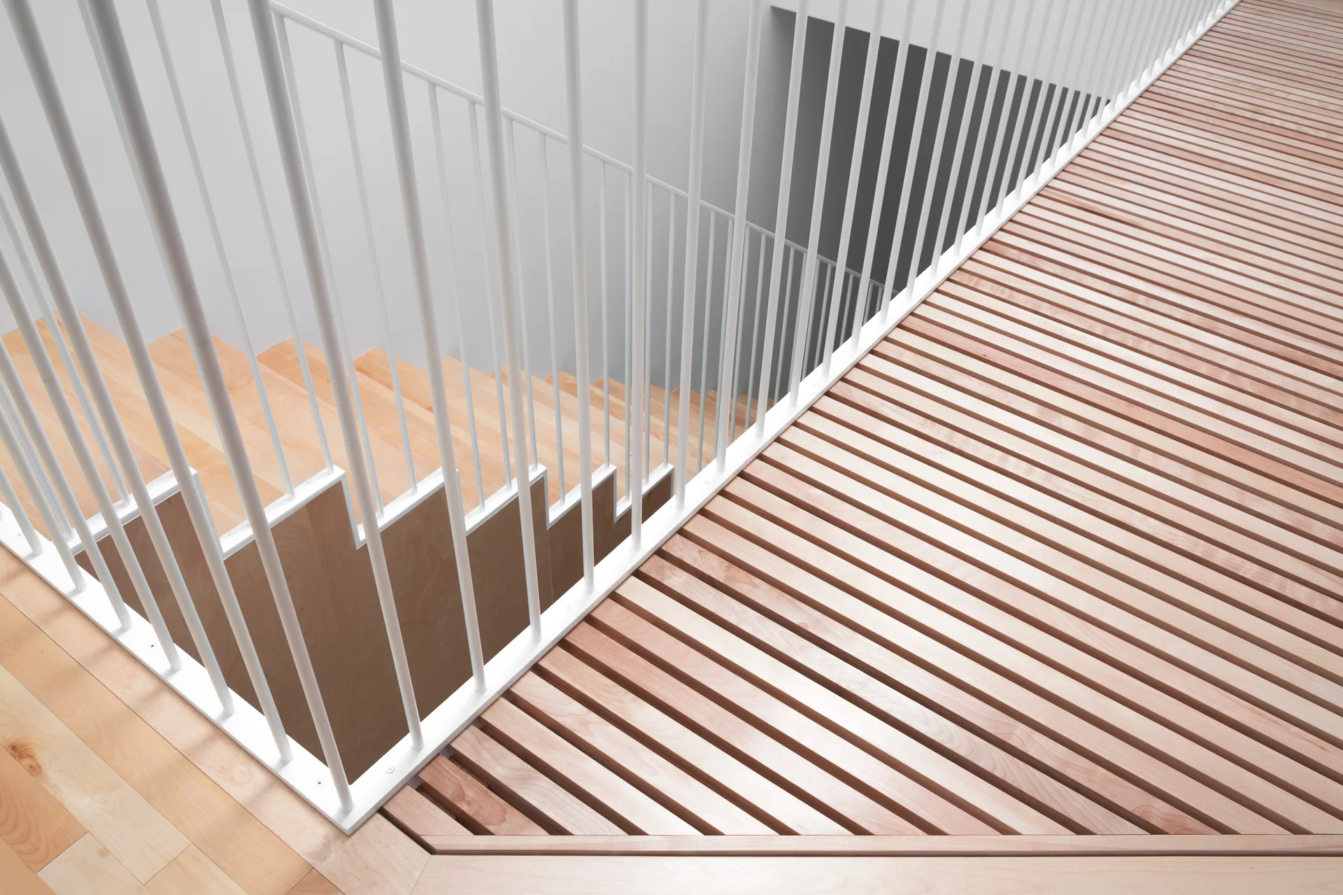 A minimalist wooden staircase with light wood treads is enclosed by a matte white vertical metal rod railing next to a slatted wood floor.