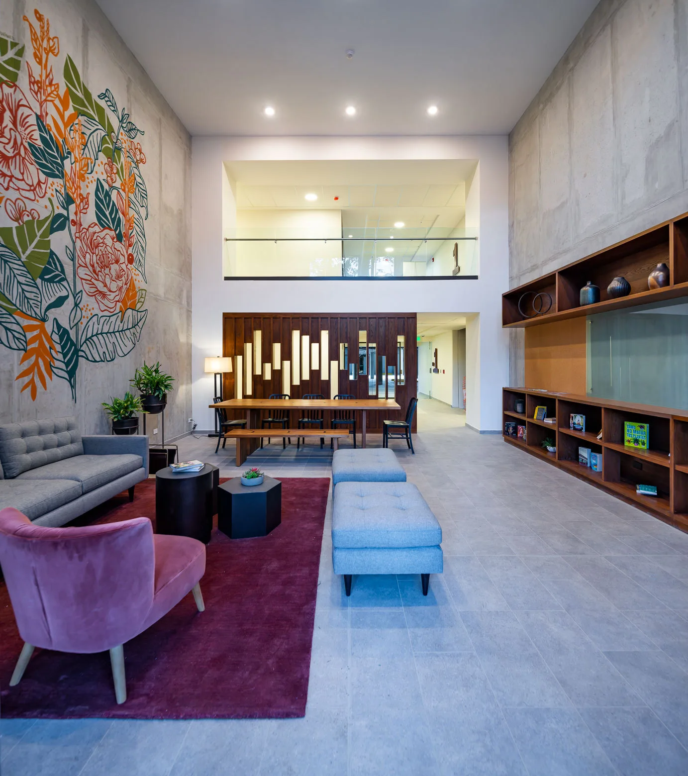 A double-height lounge features a grey sofa and pink velvet armchair on a red rug beneath an exposed concrete wall with a colorful floral mural.