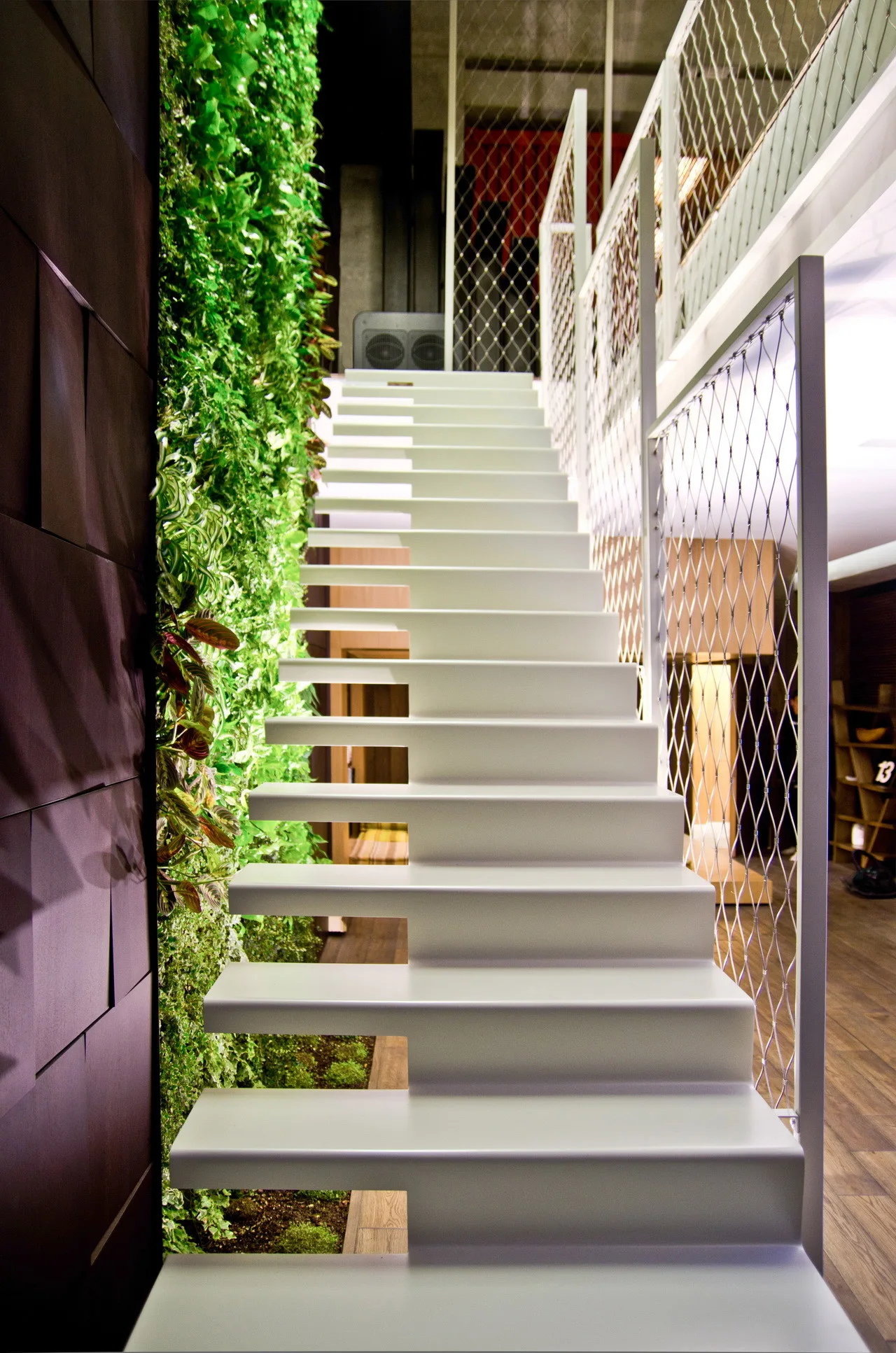A white floating staircase climbs past a tall vertical green wall and a metal wire mesh safety railing.