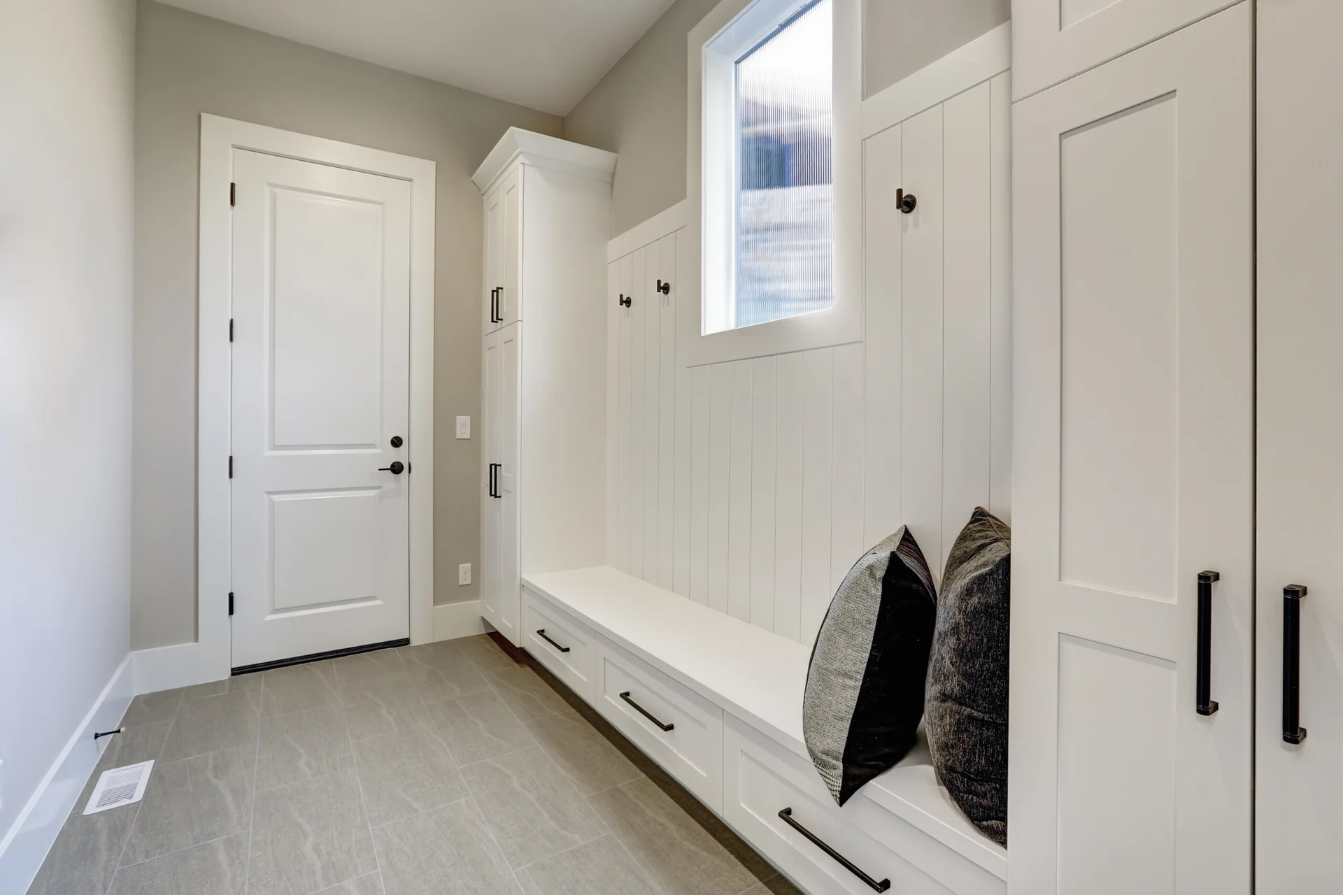 A white built-in mudroom storage bench features vertical wall paneling, tall side cabinetry, and black metal hooks on a gray tile floor.