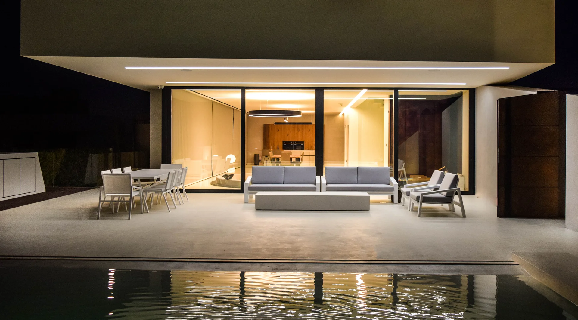 A contemporary concrete pool patio at night features minimalist gray sofas, an outdoor dining table, and a view into a warm glowing interior.