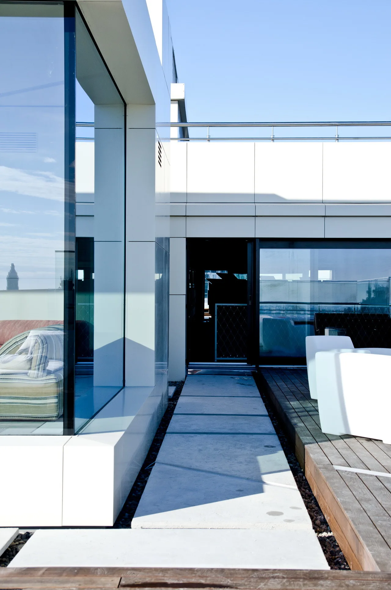 A modern rooftop terrace featuring a concrete paver walkway flanked by wooden decking, white exterior walls, and large glass windows.