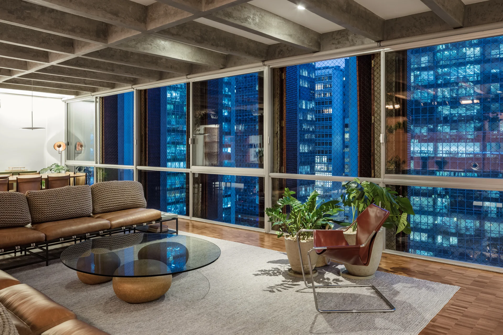 A modern living room features a tan leather sofa, glass coffee table, and large potted plants set against floor-to-ceiling windows and a concrete grid ceiling.