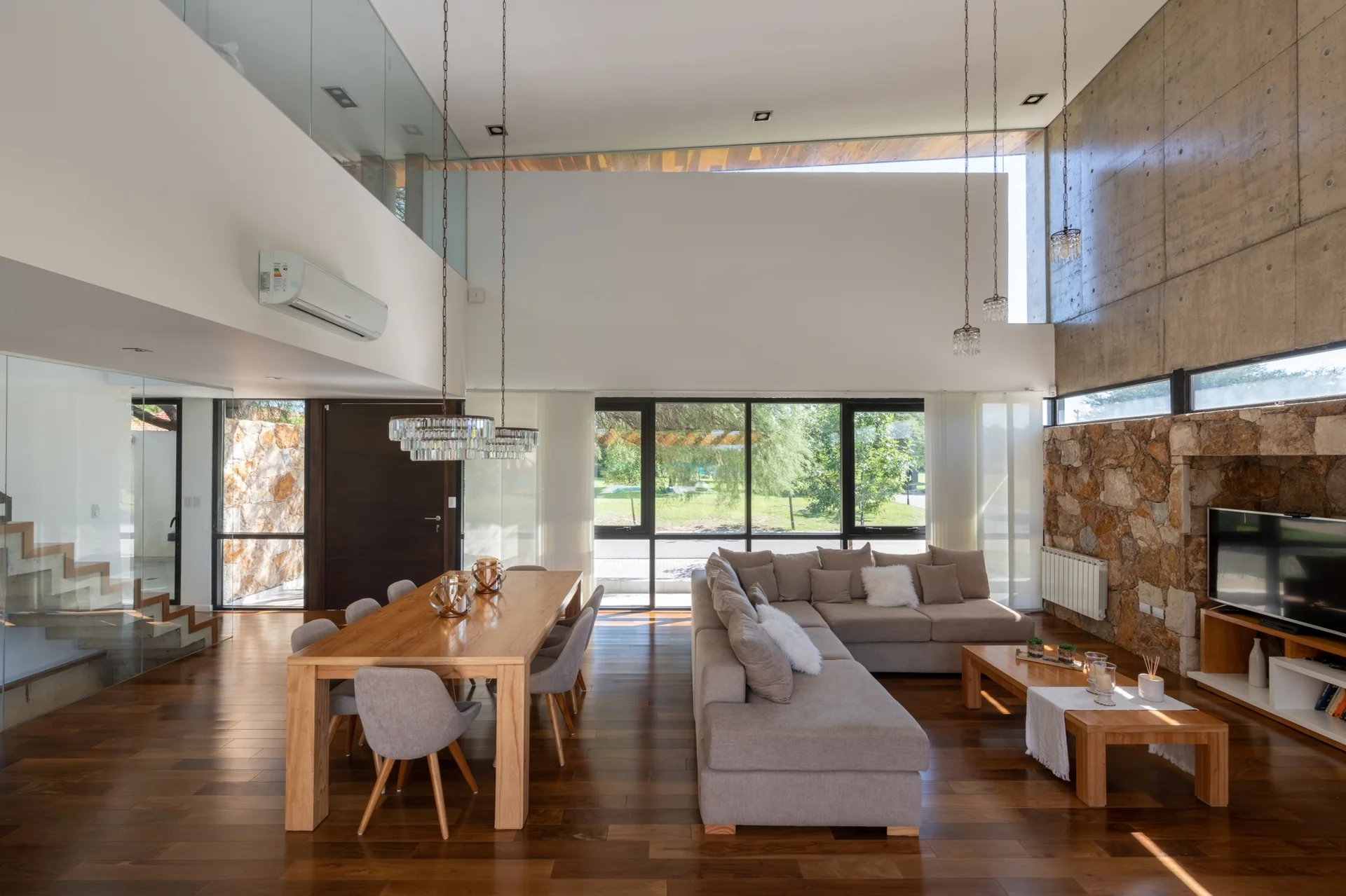 An open-plan living and dining area with a beige sectional sofa, a wooden dining table, and walls made of textured stone and exposed concrete.