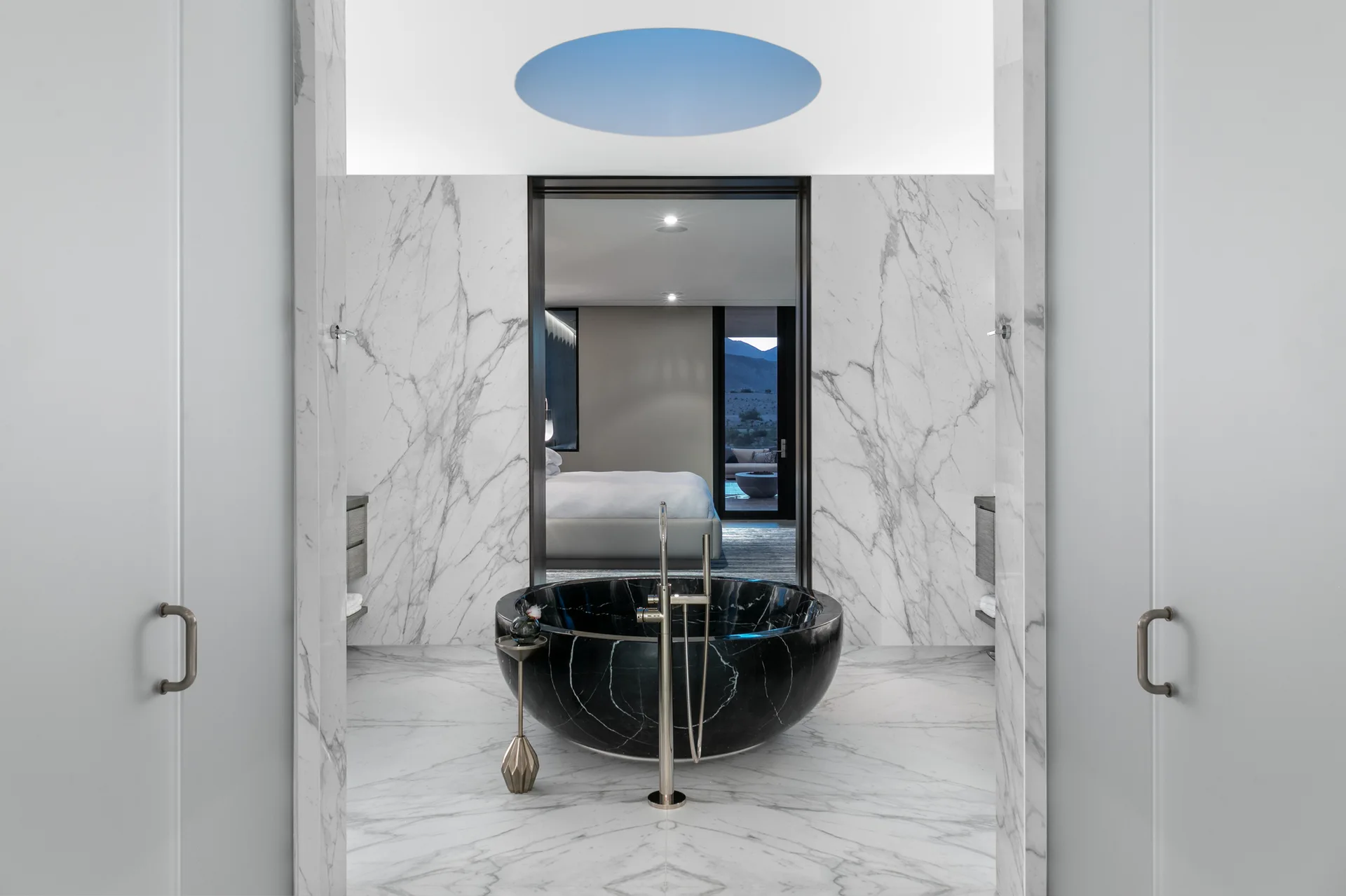 A circular black stone bathtub sits on veined white marble flooring beneath a round skylight, with a doorway looking into an adjacent bedroom.