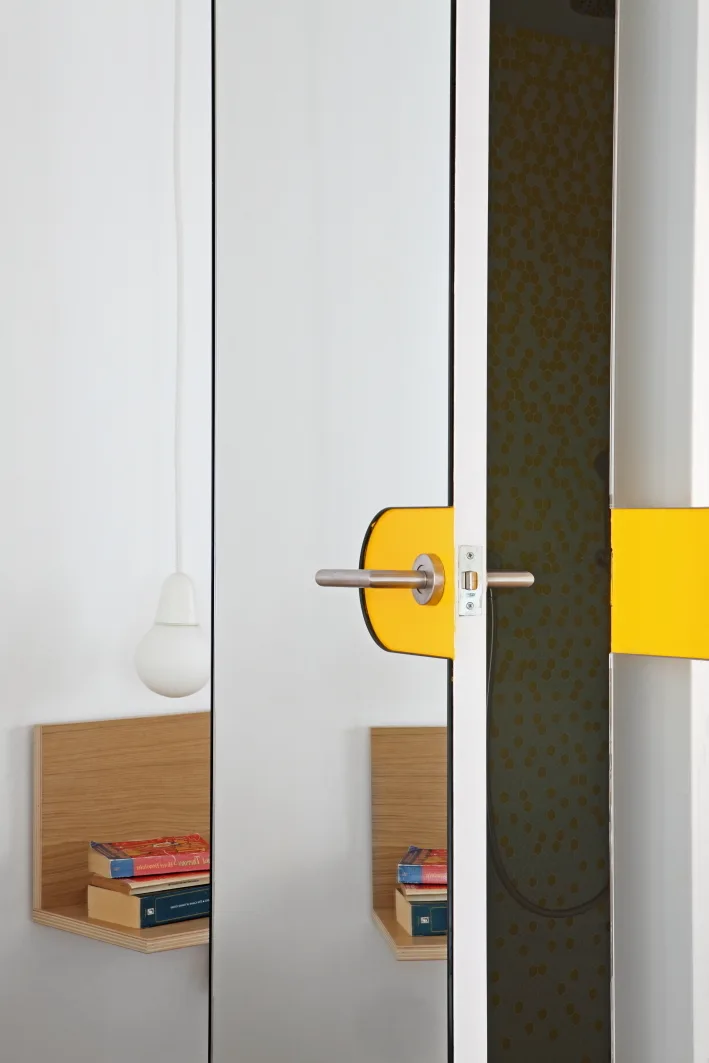 A mirrored door with a yellow accent plate reflects a wood floating shelf and reveals yellow hexagonal tiles in the background.