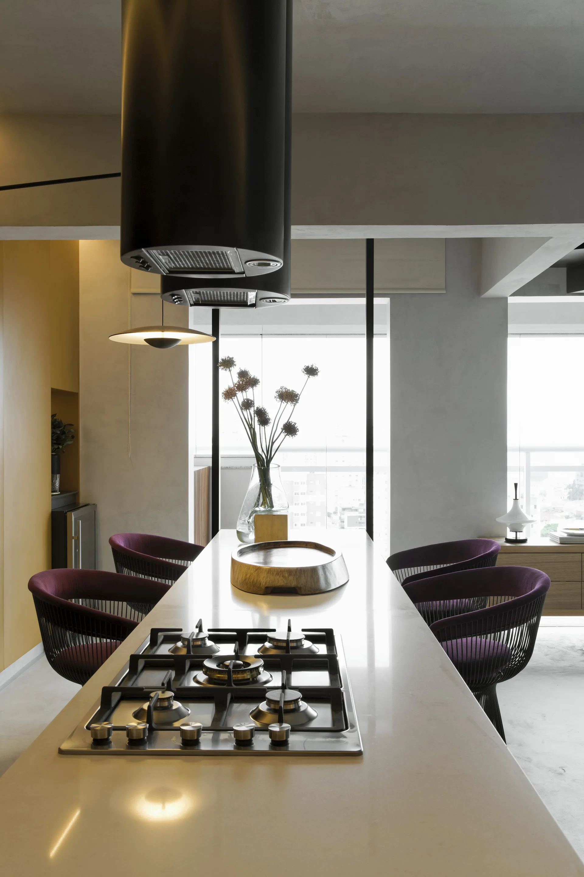 A long kitchen island with an integrated gas stovetop is paired with purple barrel chairs under cylindrical black range hoods.
