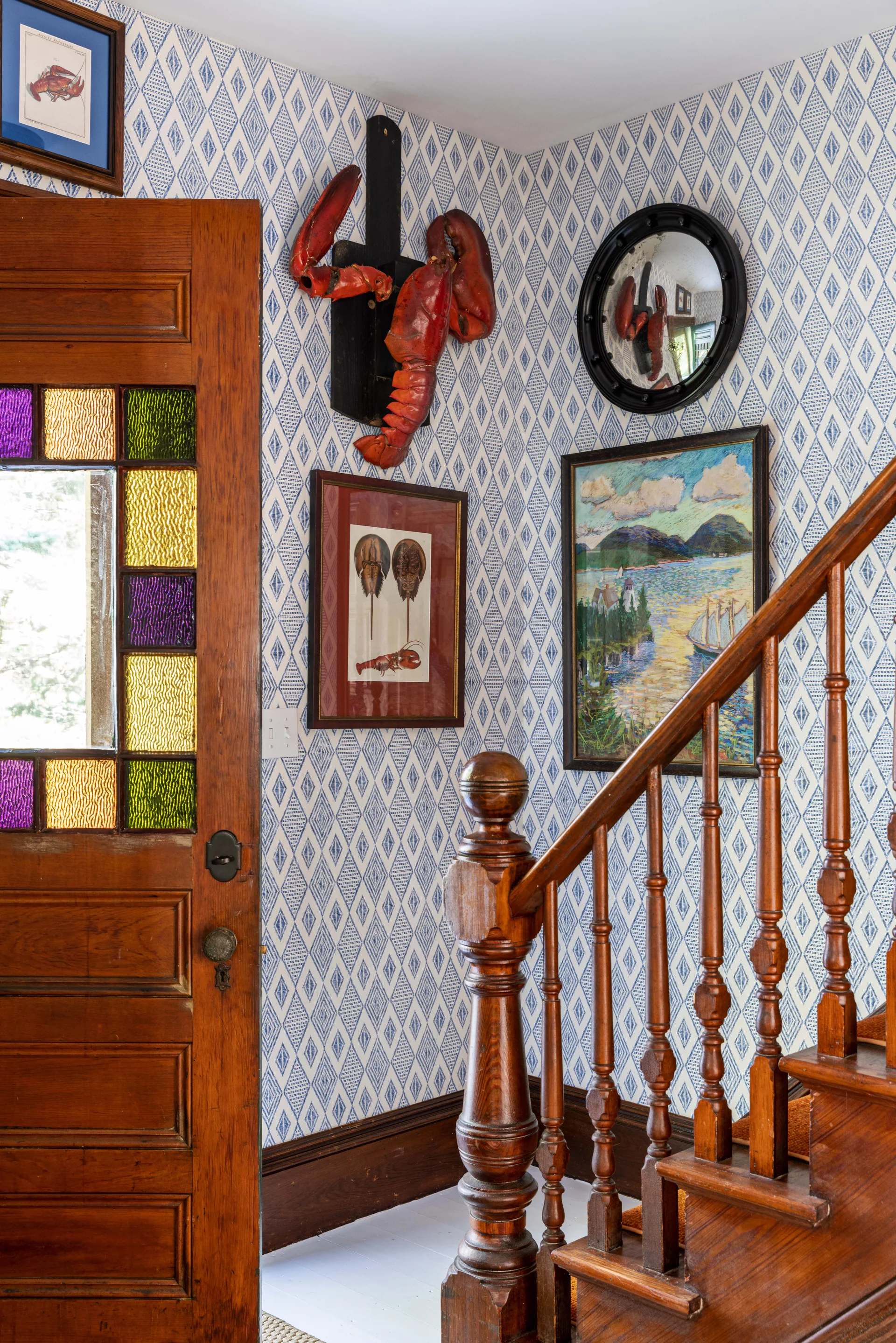 A wooden staircase and door with stained glass stand against walls covered in blue and white diamond-patterned wallpaper decorated with coastal art.
