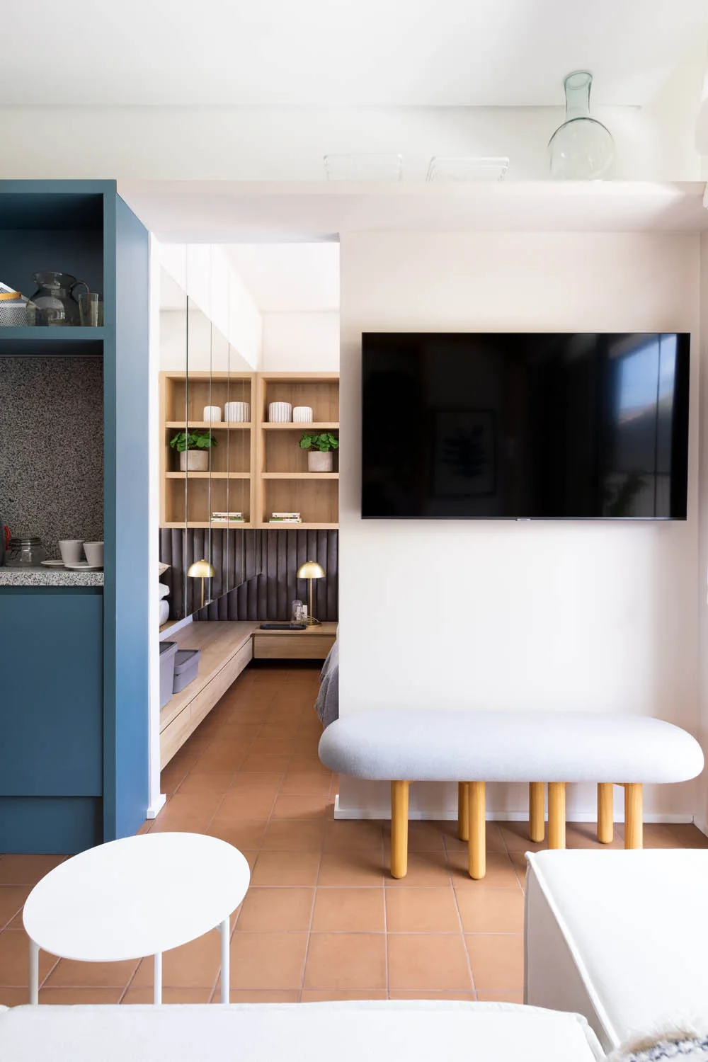 A modern living area with a grey upholstered bench, a wall-mounted TV, terracotta floor tiles, and a view into a room with wood shelving and mirrors.