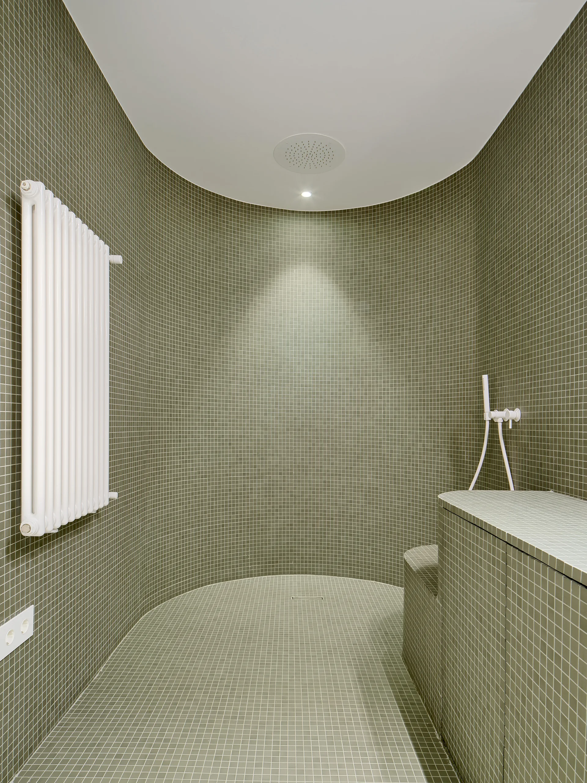 A curved walk-in shower features sage green mosaic tiles, a built-in tiled bench, and a white vertical radiator.