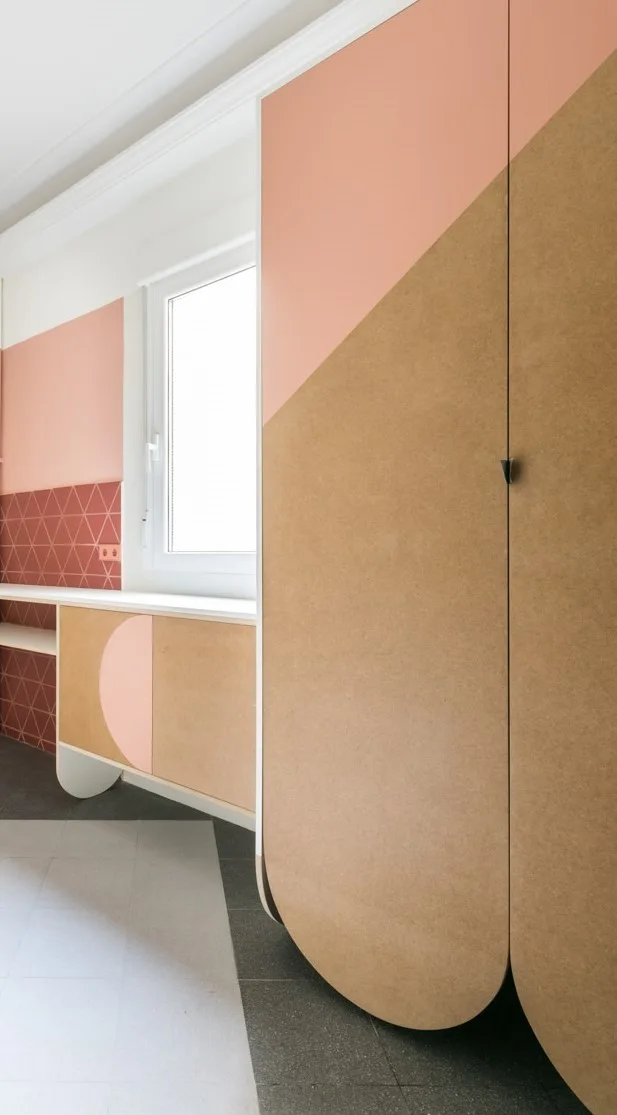 Custom peach and tan color-blocked wardrobe doors with rounded bottoms stand next to a built-in desk area with geometric pink wall tiles.