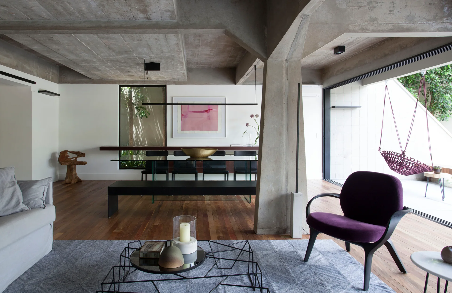 A contemporary open-plan living and dining area featuring a raw concrete column and ceiling, dark wood floors, and a purple accent chair.