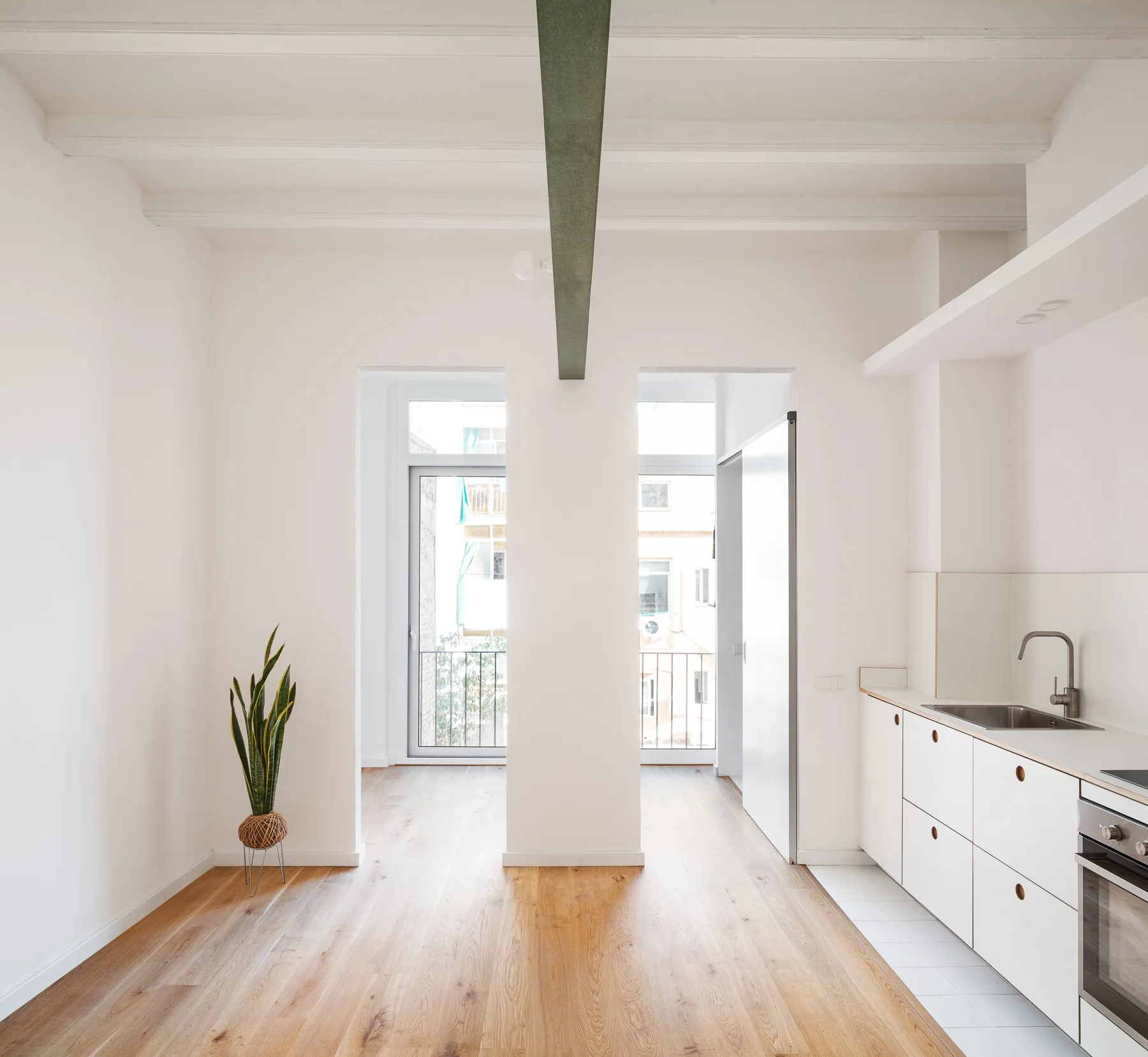 A minimalist white kitchen and living space features wood floors, dual symmetrical doorways, and a single green beam on a white beamed ceiling.