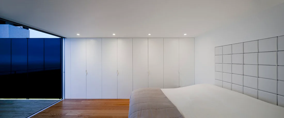 A minimalist white bedroom featuring a wall of built-in floor-to-ceiling closets, a bed with a square-tufted headboard, and wood flooring.
