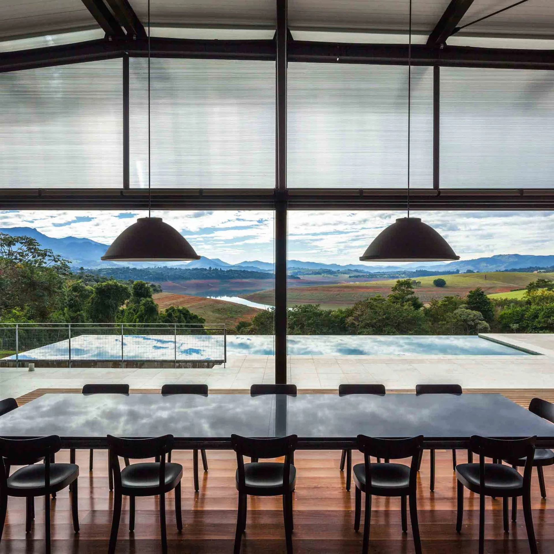 A long black dining table with black chairs sits in front of a floor-to-ceiling glass wall overlooking an infinity pool and hills.