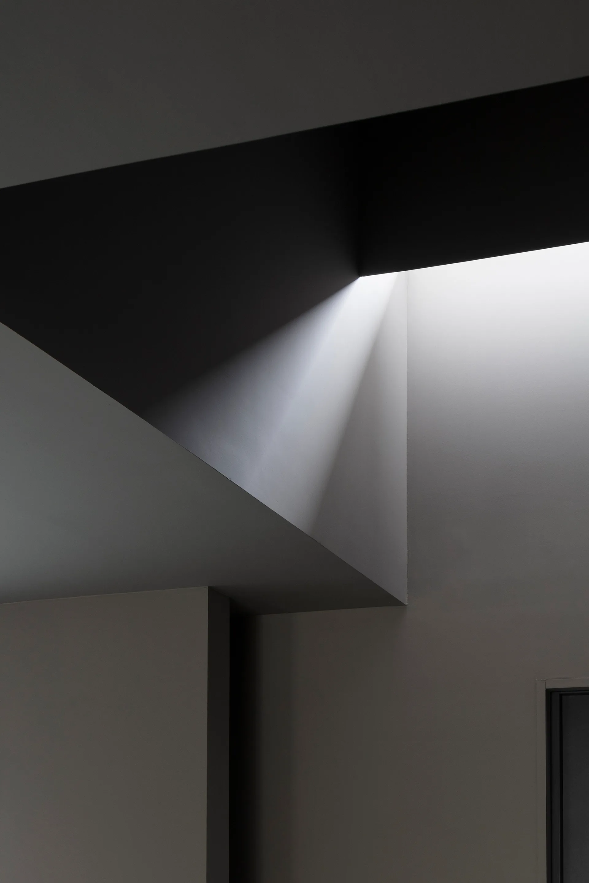 Sharp geometric ceiling planes and smooth grey walls frame a bright beam of light from a hidden overhead opening.