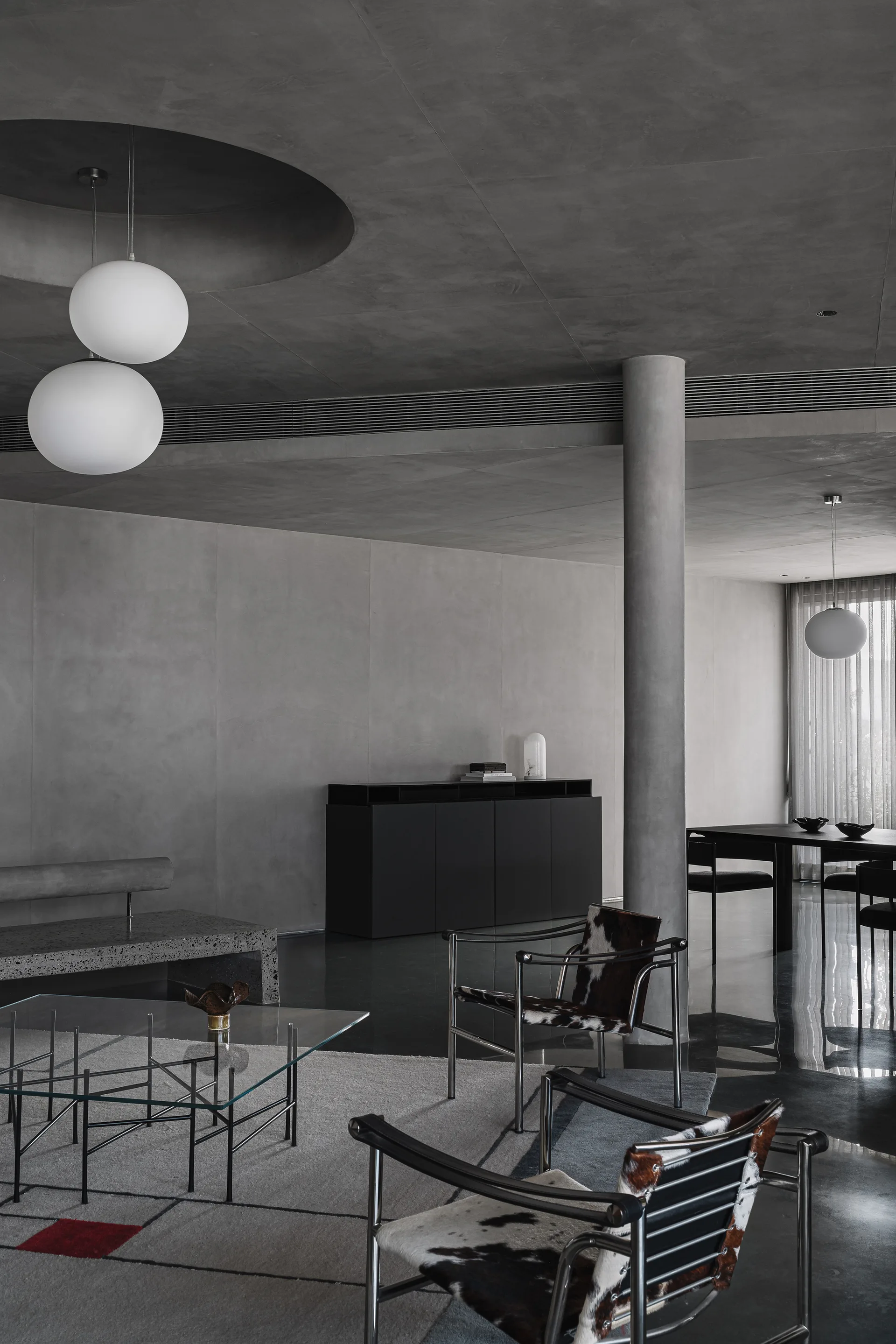 A minimalist gray concrete living room features two cowhide and chrome lounge chairs, a glass coffee table, and spherical white pendant lights.