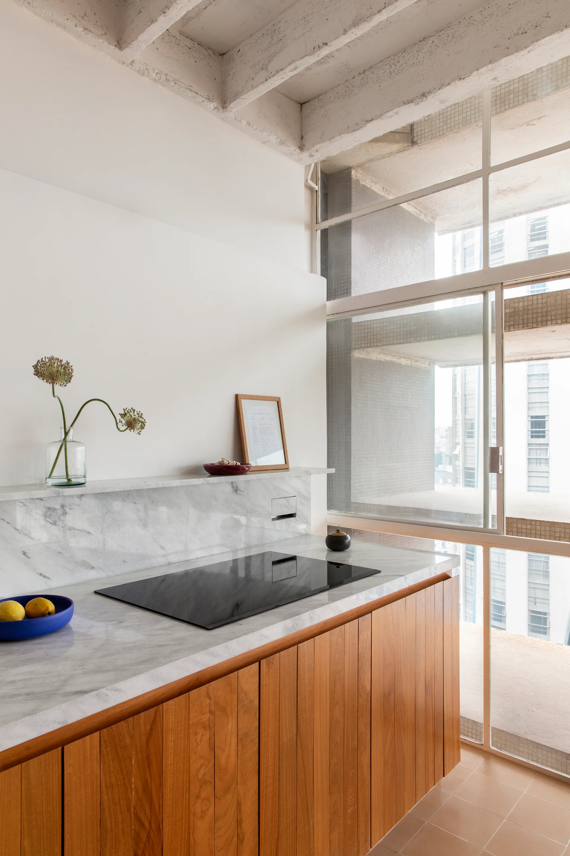 A modern kitchen featuring vertical wood base cabinets, a white marble countertop with an integrated cooktop, and a large sliding glass window.