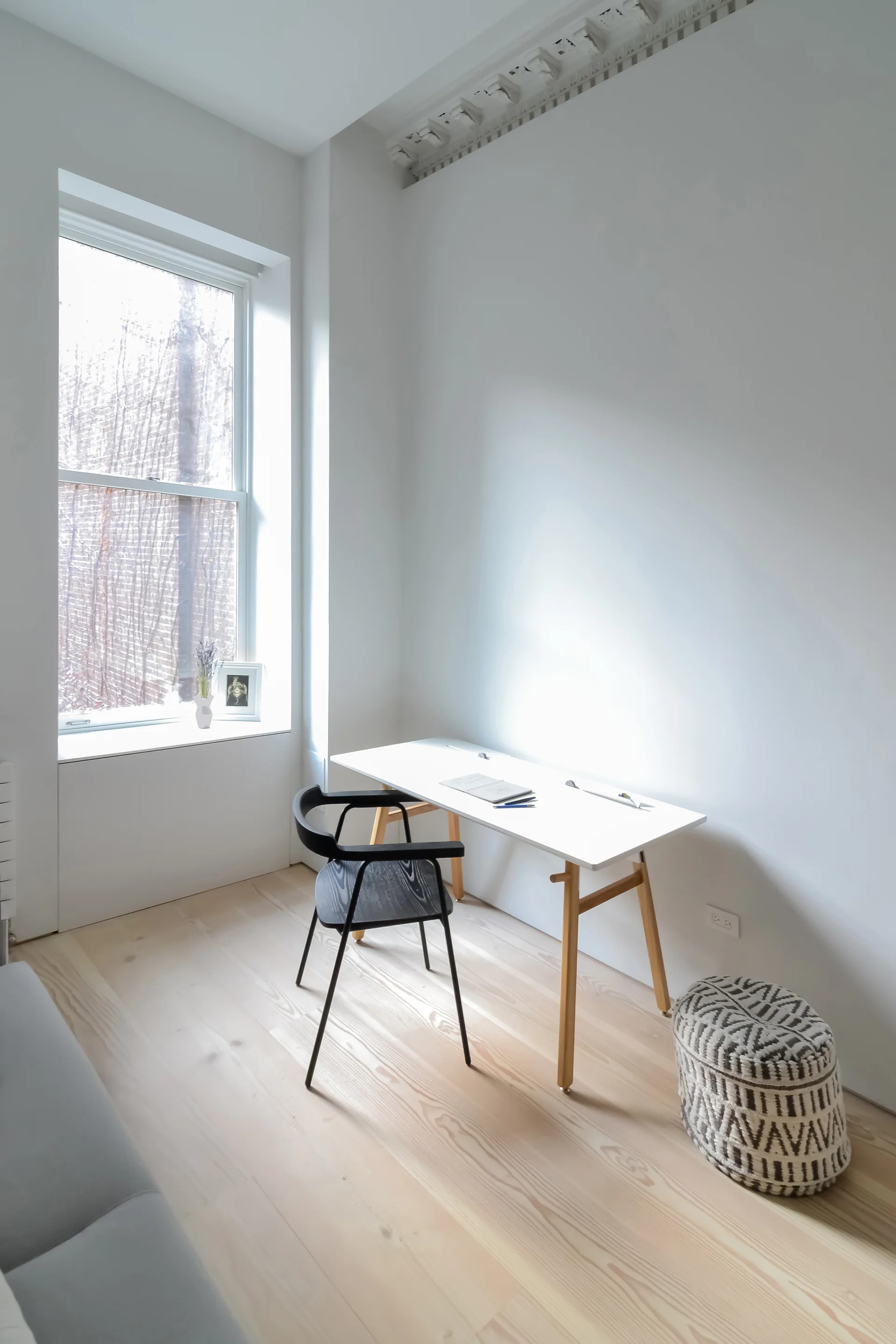 A minimalist home office features a white desk, a black wooden chair, and a patterned ottoman on light wood floors.