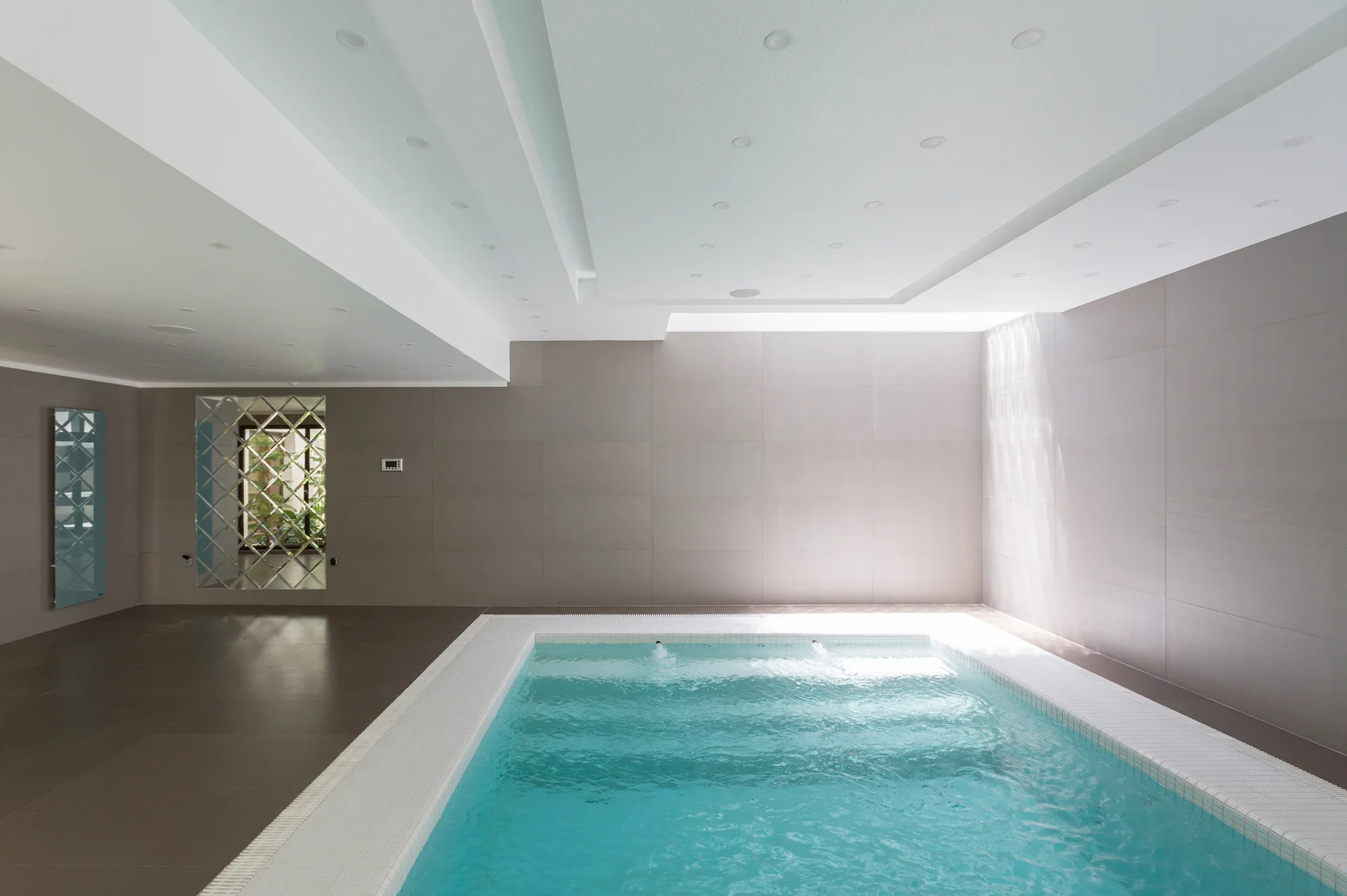 A sunken indoor swimming pool with turquoise water is surrounded by matte grey wall tiles and a multi-tiered white ceiling with recessed spotlights.