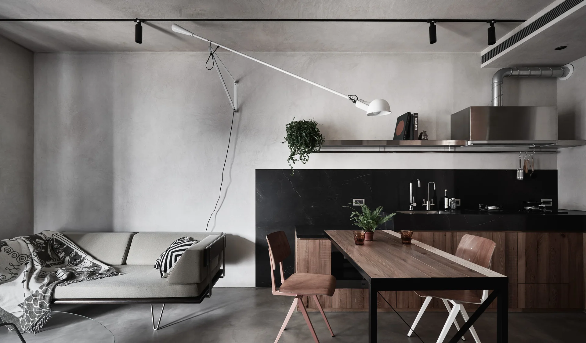 A modern open-plan industrial kitchen and living area features concrete walls, black backsplash, wooden cabinetry, and a low grey sofa.
