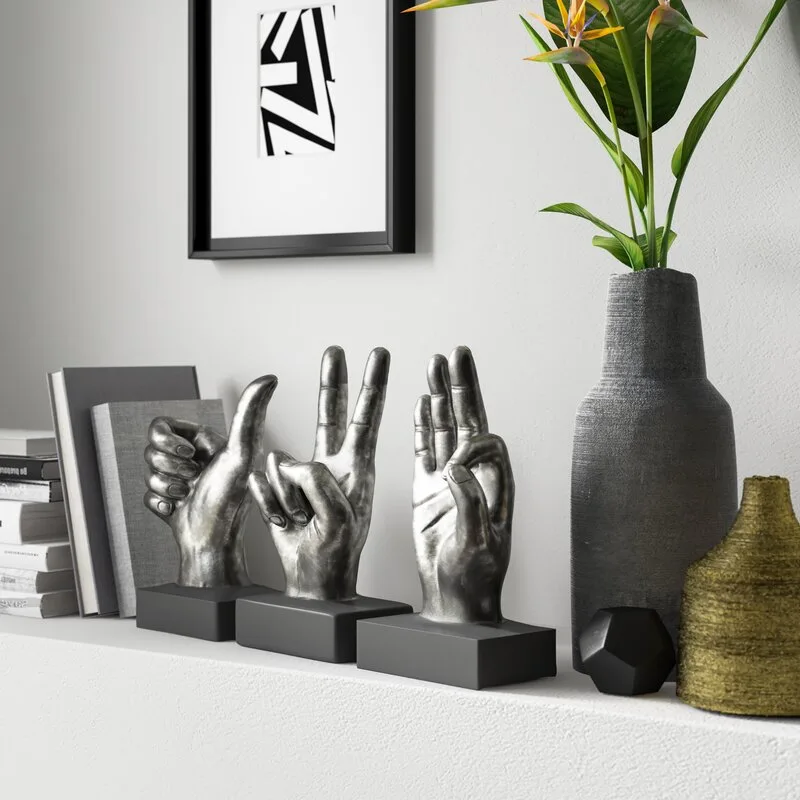 Three silver hand gesture sculptures sit on a white shelf next to books, a tall grey textured vase, and a small gold vase.