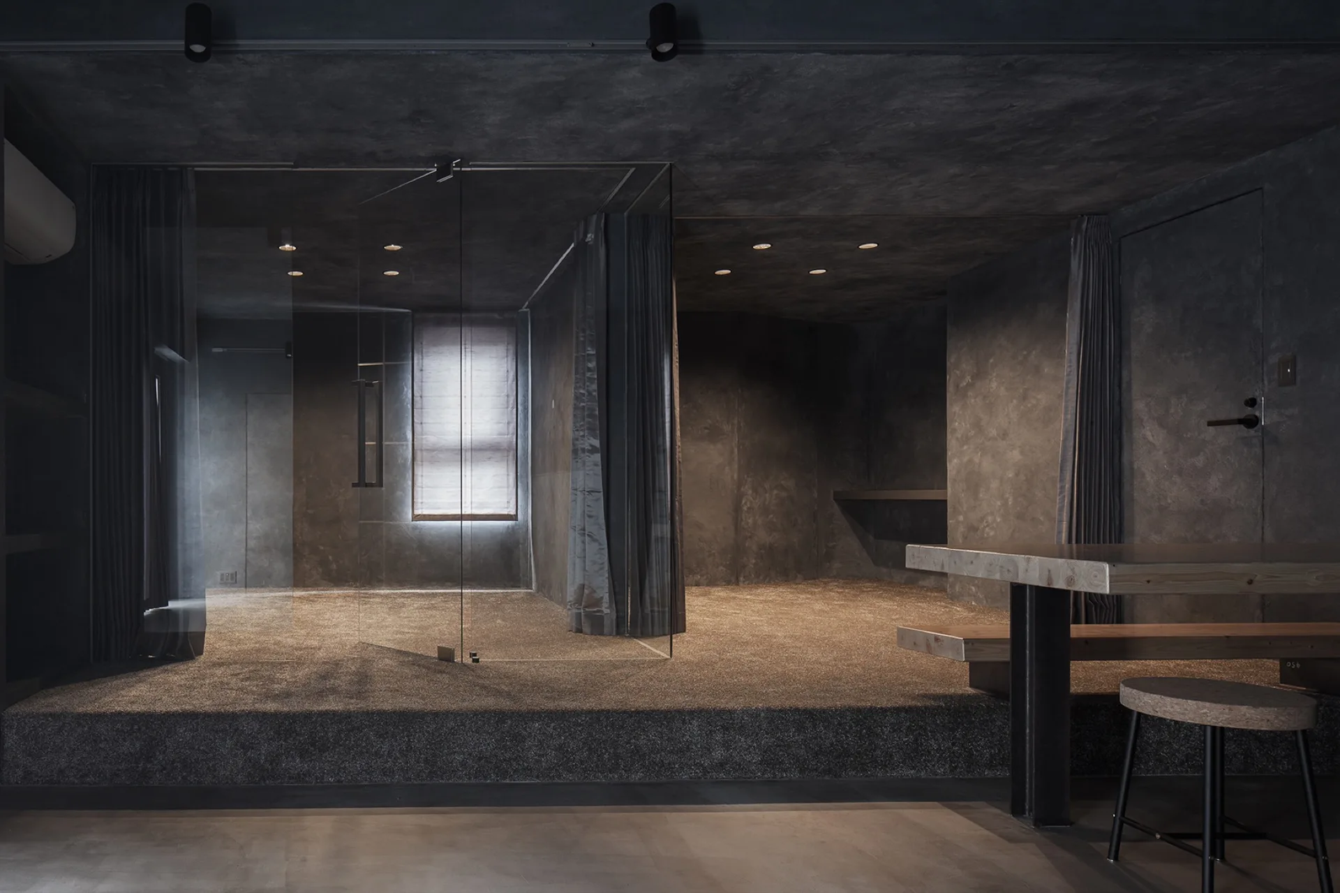 A dark gray minimalist interior features a raised carpeted platform divided by a frameless glass partition and a wooden table in the foreground.
