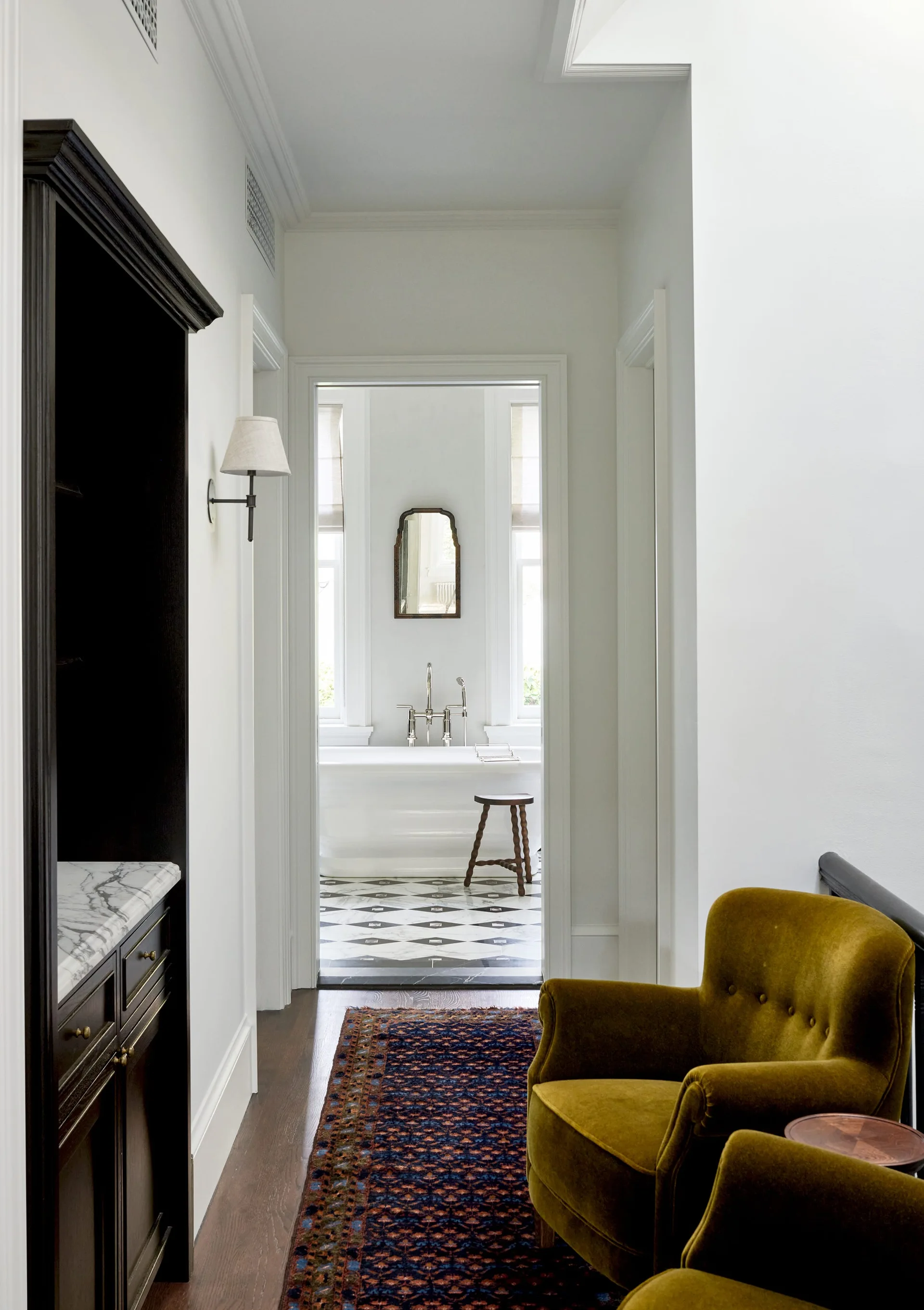 A hallway with two olive velvet chairs leads to a bright bathroom with a soaking tub and diamond pattern tile floor.