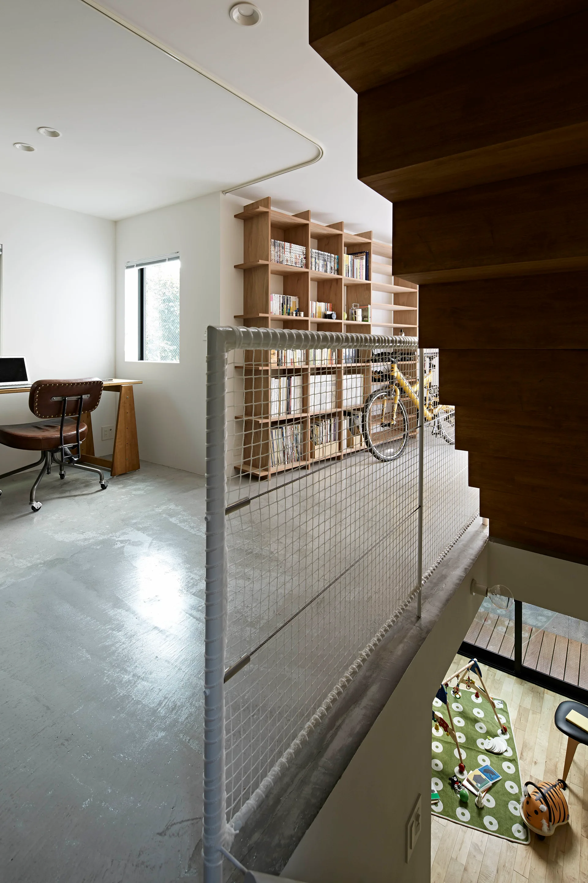 A split-level landing features concrete flooring, a white mesh safety railing, a wood desk, and a large wooden bookshelf.