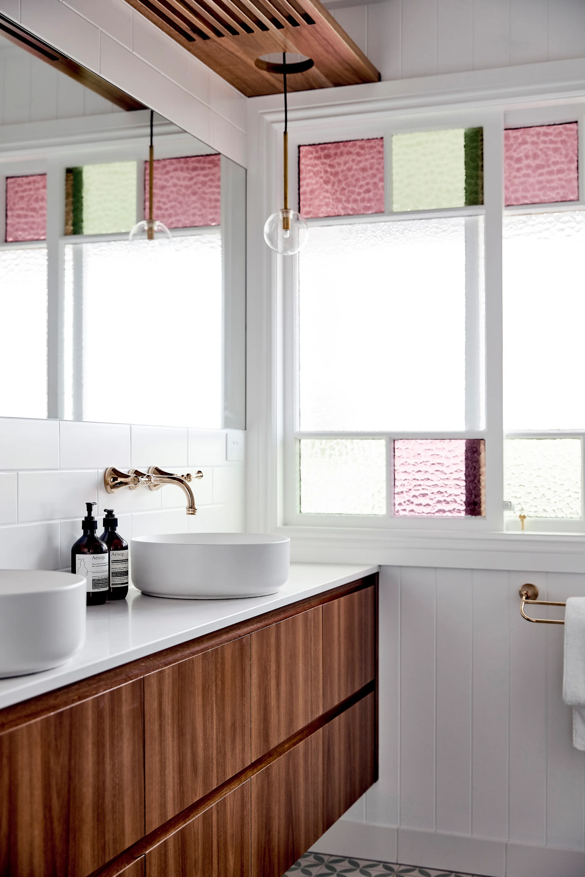 A modern bathroom features a floating wood vanity with white vessel sinks, gold hardware, and windows with pink and green stained glass inserts.