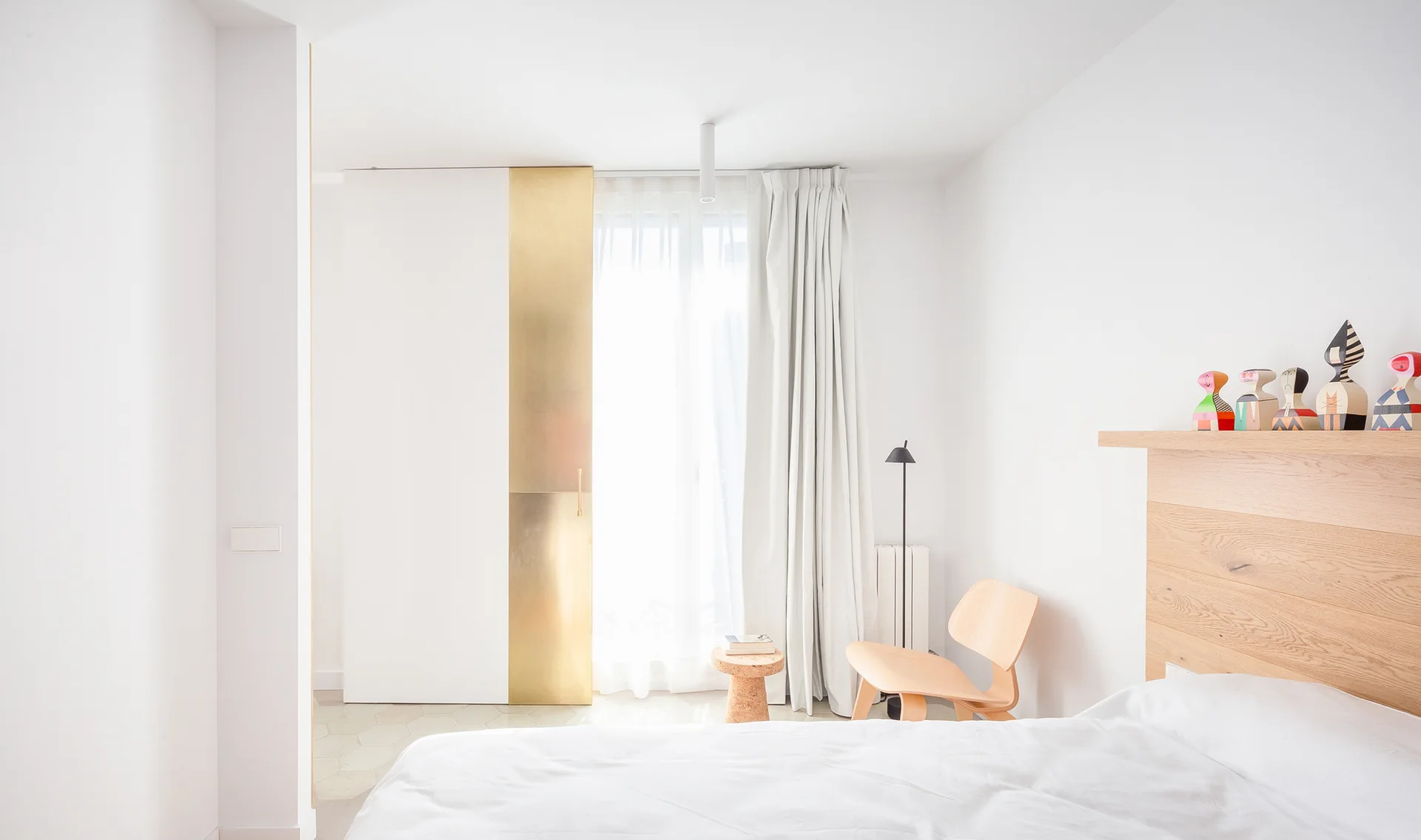 A minimalist white bedroom with a light wood headboard, a molded plywood chair, and a sliding door featuring a polished brass vertical stripe.