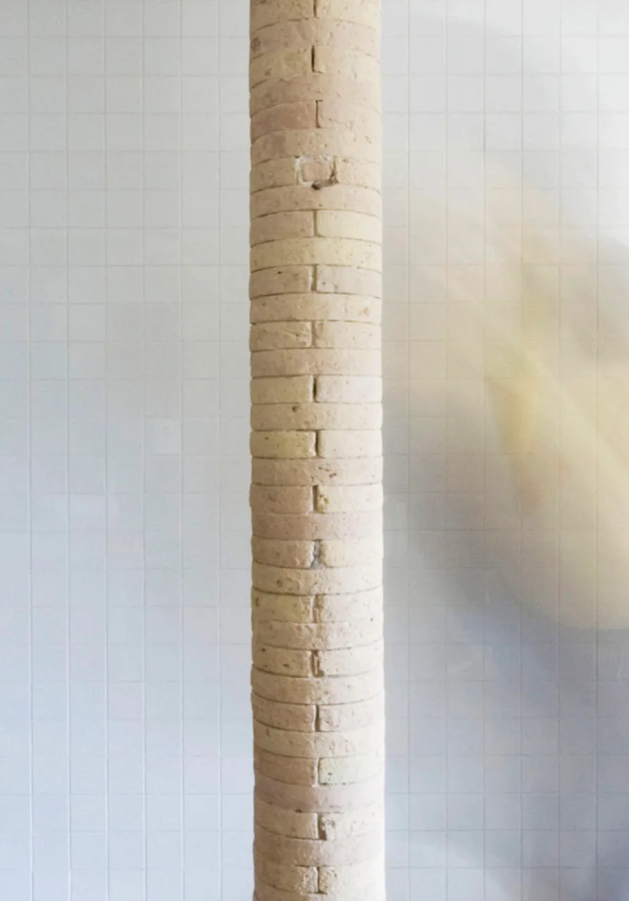 A cylindrical column constructed of stacked beige bricks stands in front of a white grid-tiled wall.