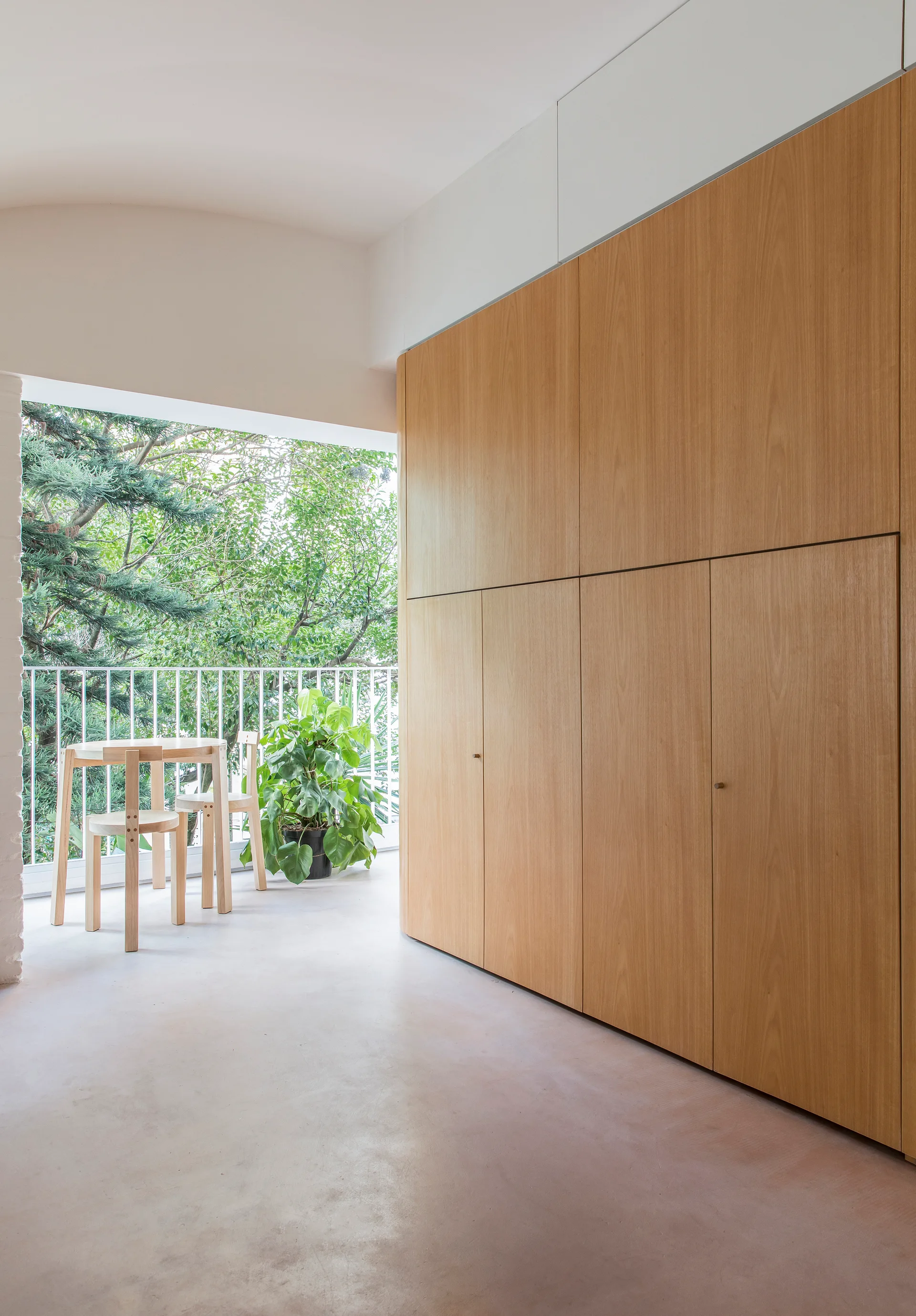 A minimalist room with floor-to-ceiling light wood cabinets that open onto a bright balcony with a small wooden table and chairs.