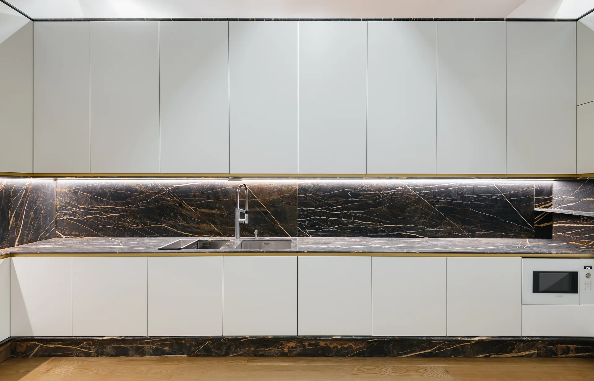 A modern kitchen with upper and lower white handleless cabinets contrasted by a dark marble countertop and backsplash with gold veining.
