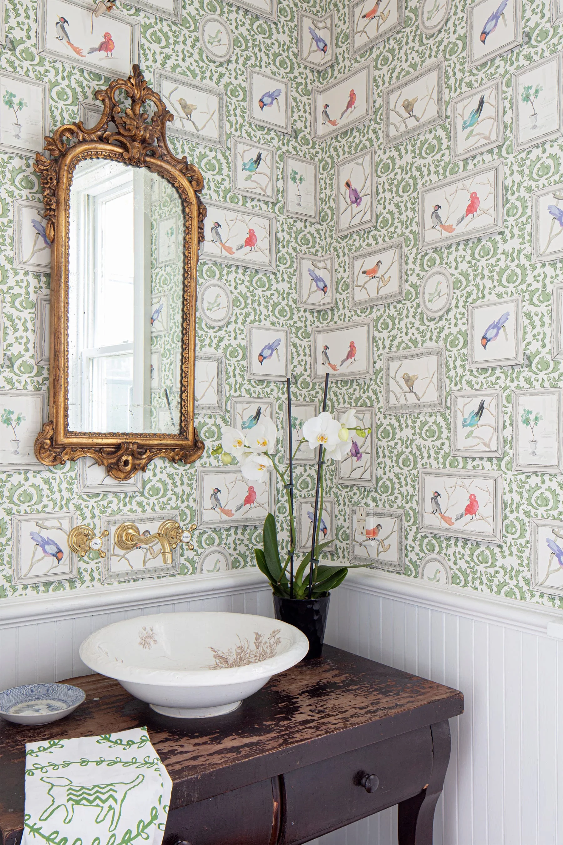 A powder room features green and white bird wallpaper, white beadboard wainscoting, and a distressed dark wood vanity with a white ceramic vessel sink.