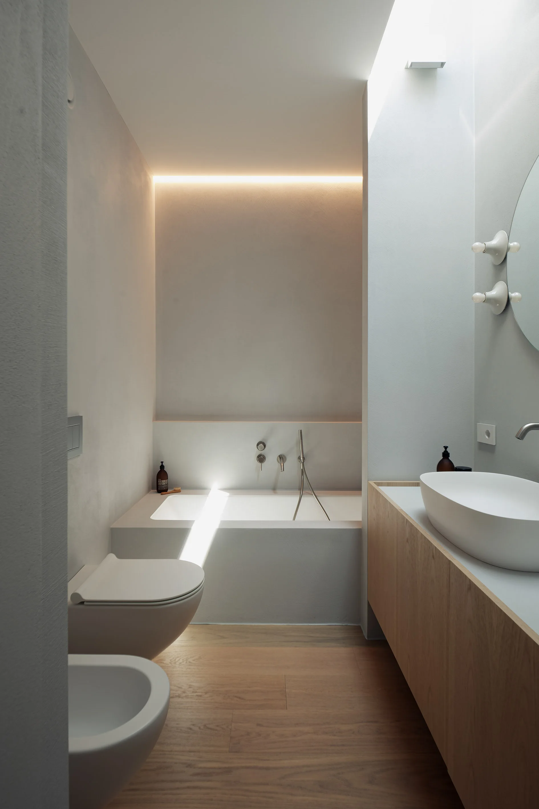 A minimalist modern bathroom featuring a built-in bathtub, warm cove lighting, a wall-mounted wood vanity with a vessel sink, and a skylight.