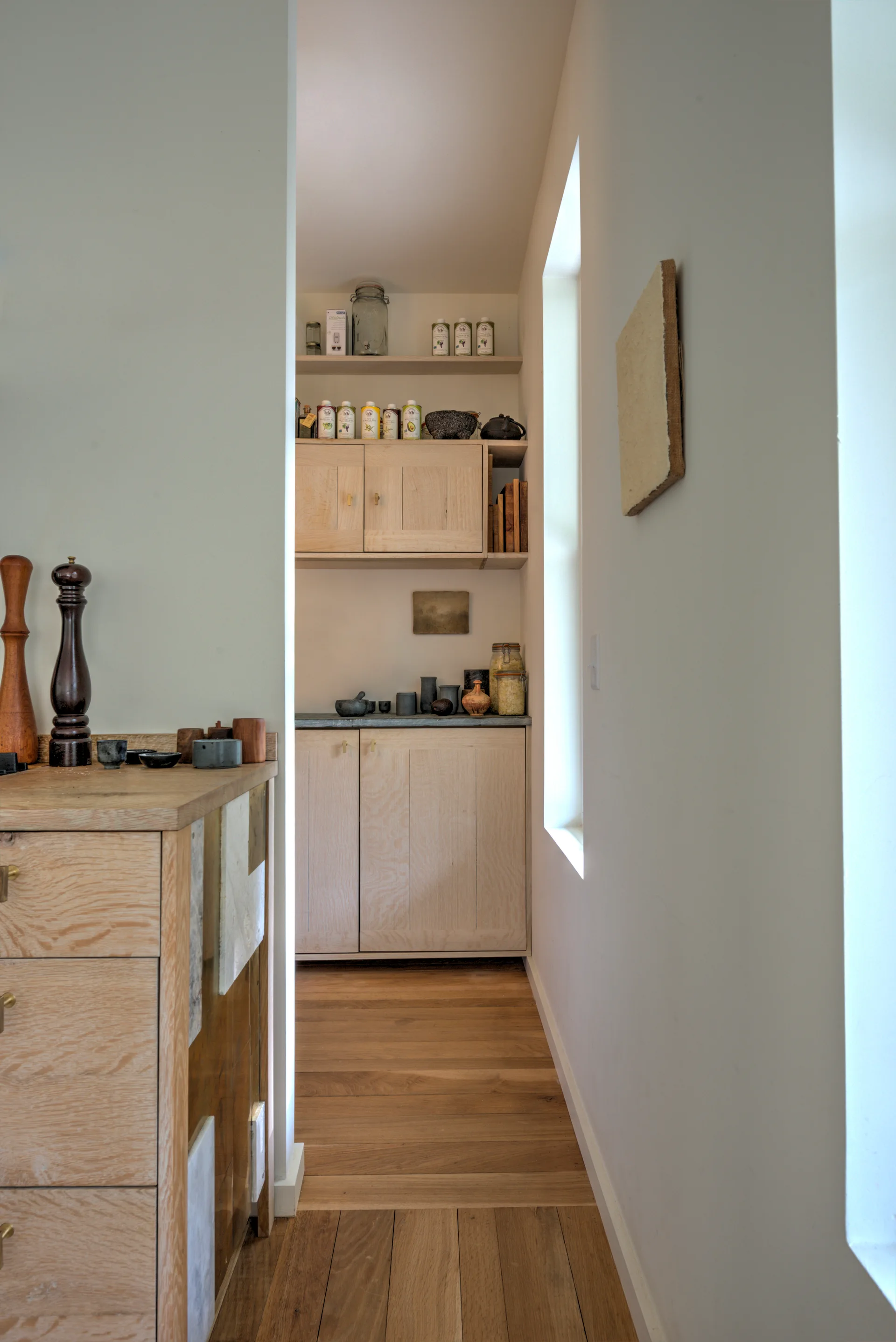 A narrow kitchen corridor features light oak cabinets, a hardwood floor, and a tall narrow window casting natural light on the workspace.