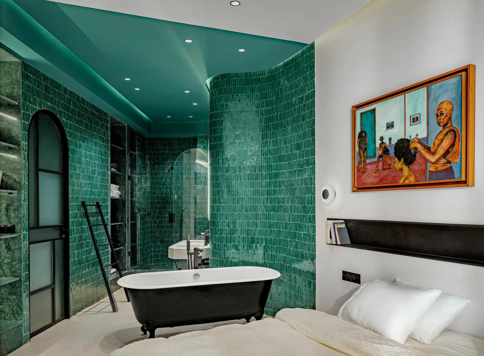 An open concept bedroom and bathroom suite with a black freestanding bathtub against a curved wall of glossy green tiles.