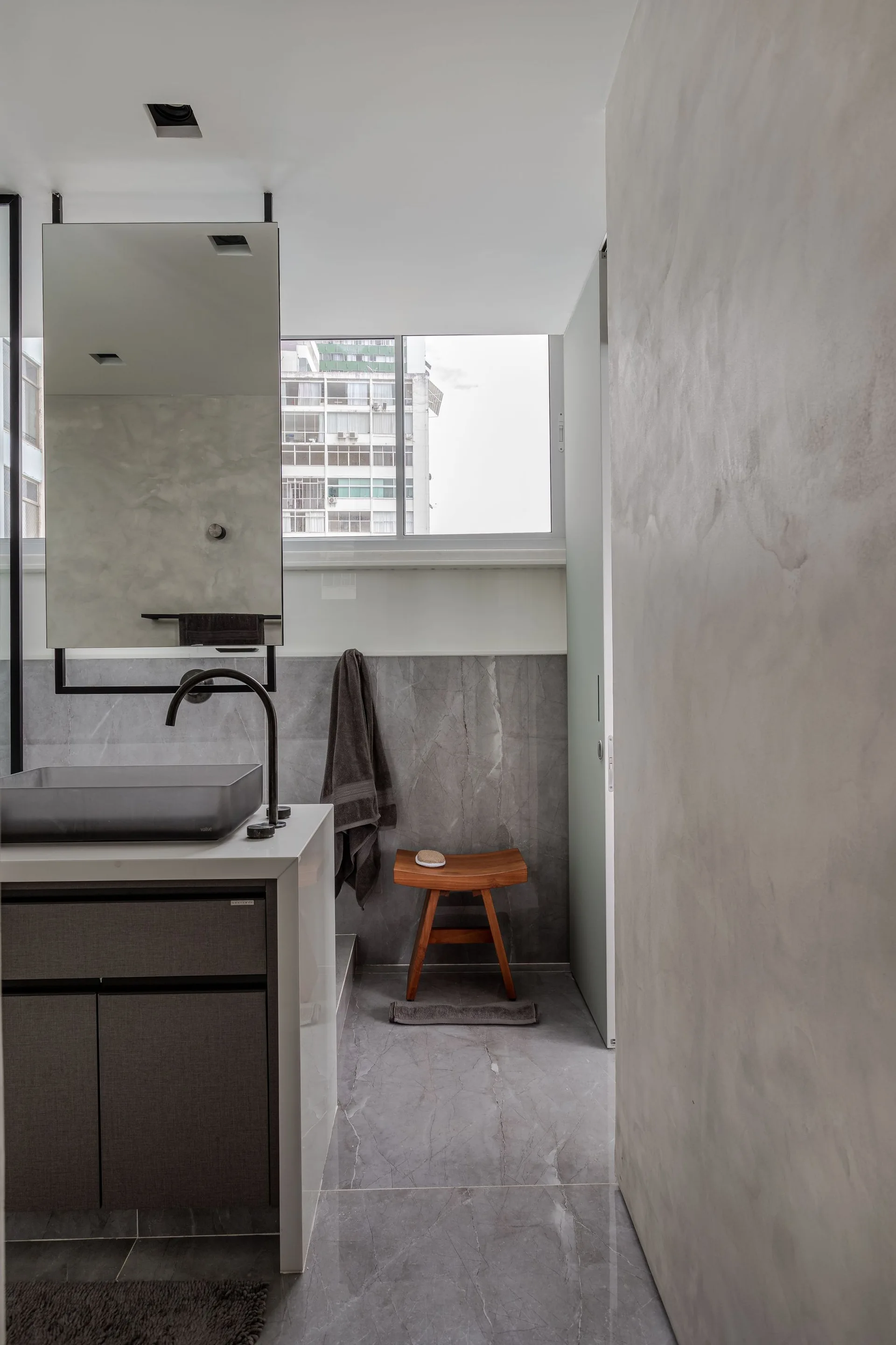 A modern grey bathroom featuring a vanity with a dark basin, a suspended rectangular mirror, a wooden stool, and large grey stone tiles.