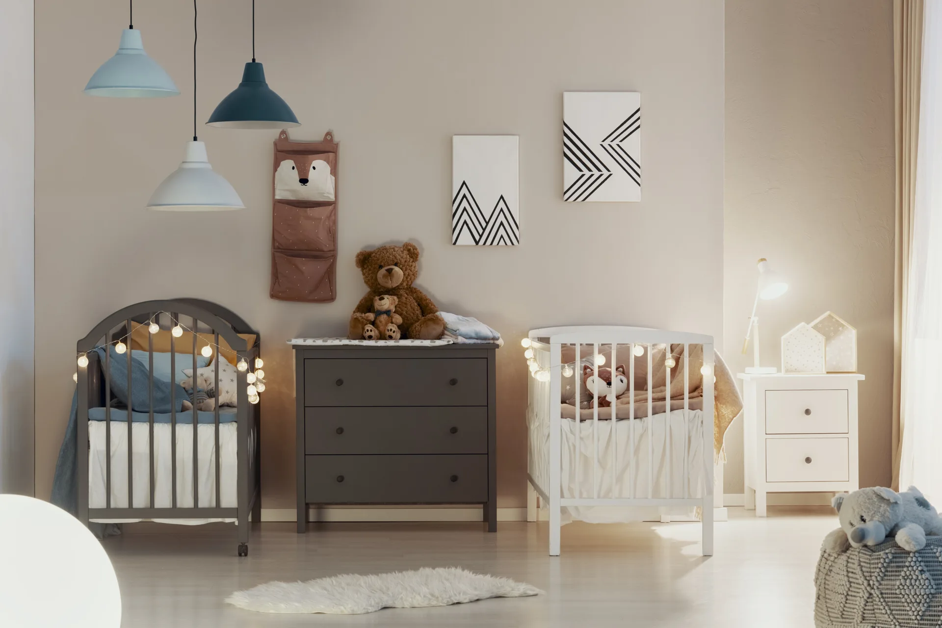 A twin nursery features a grey crib and a white crib flanking a dark grey dresser under three hanging pendant lights.