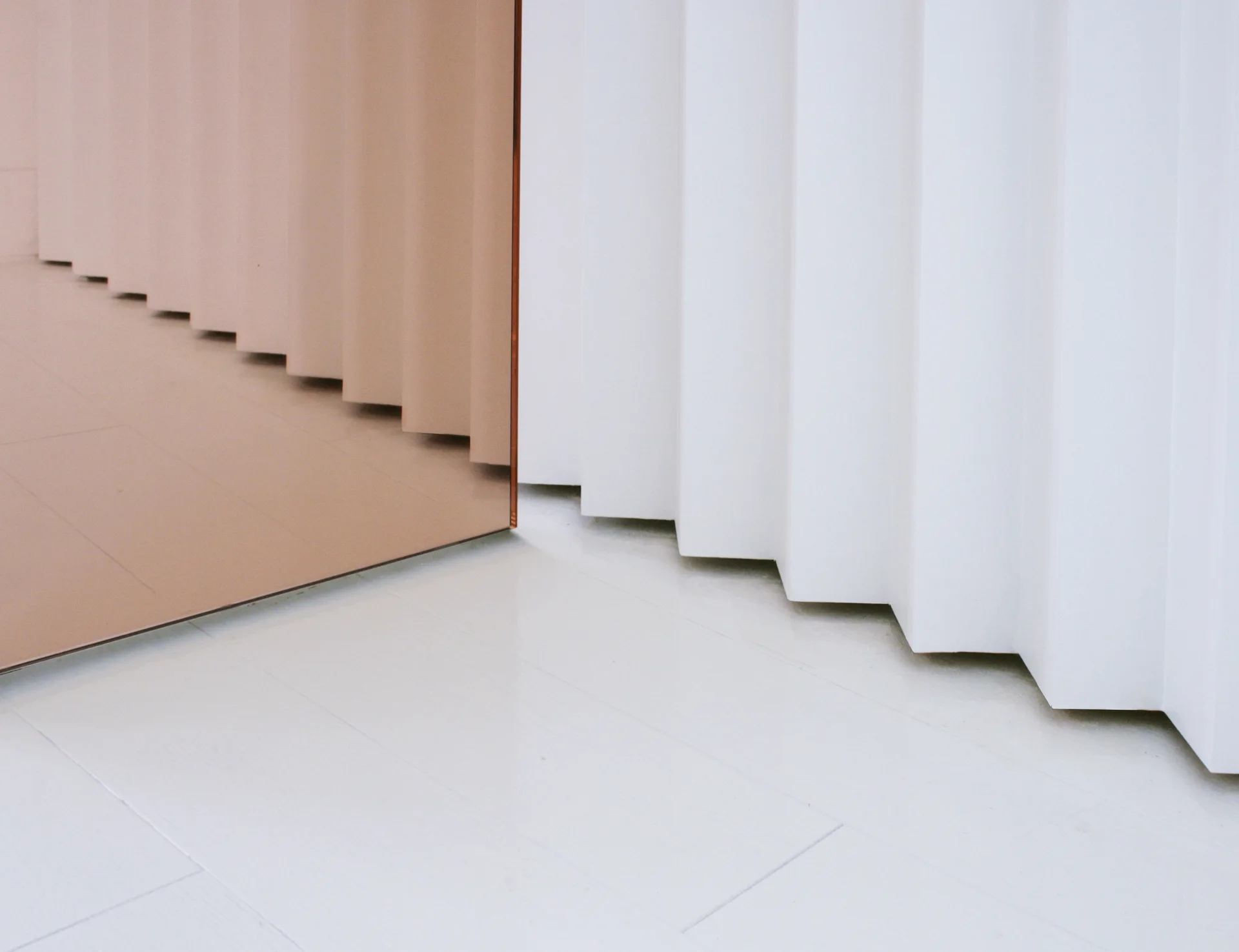A white pleated wall panel reflects in an adjacent bronze tinted mirror on a smooth white floor.