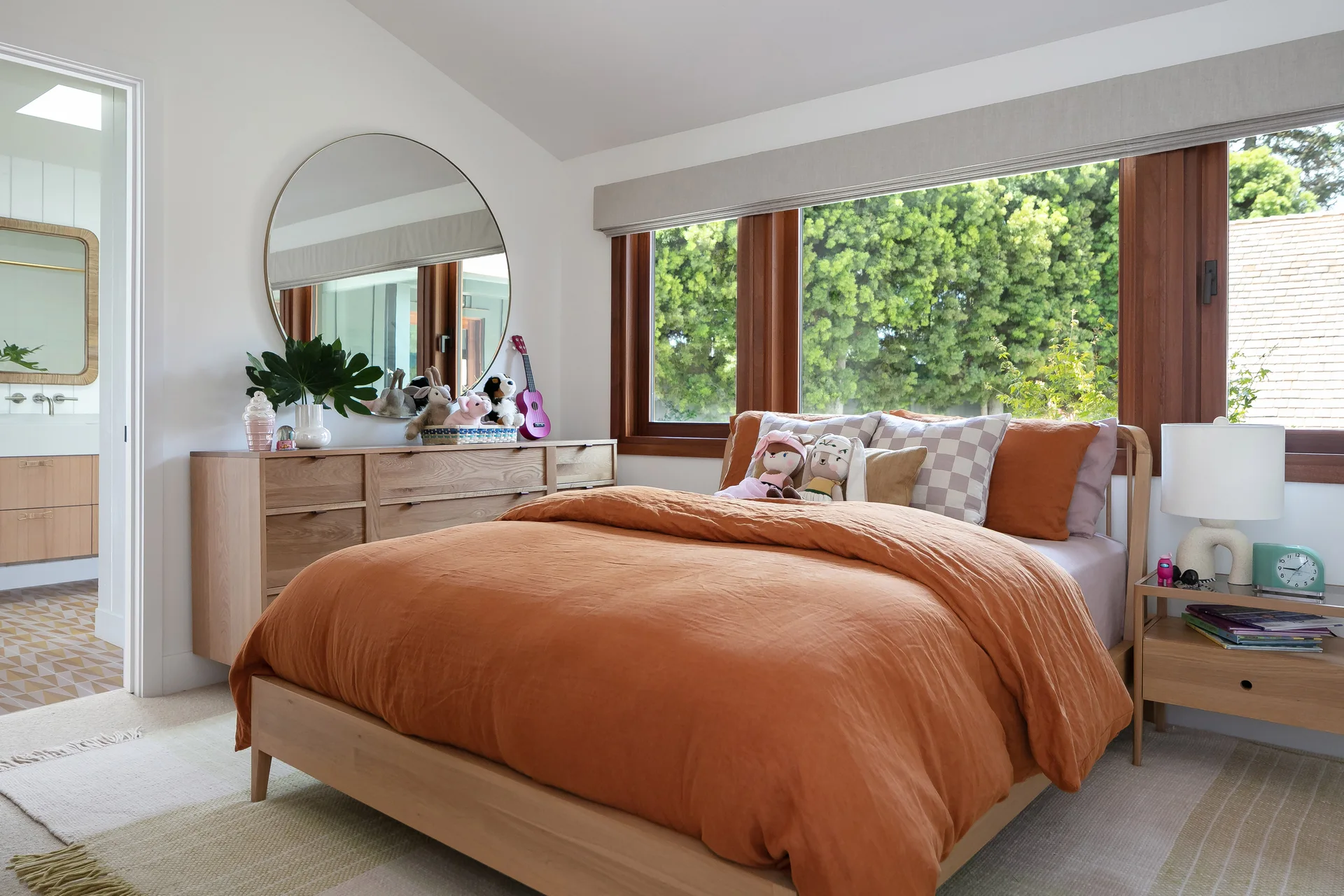A modern bedroom features a light wood bed with terracotta bedding, a large round mirror above an oak dresser, and a wide wood-trimmed window.