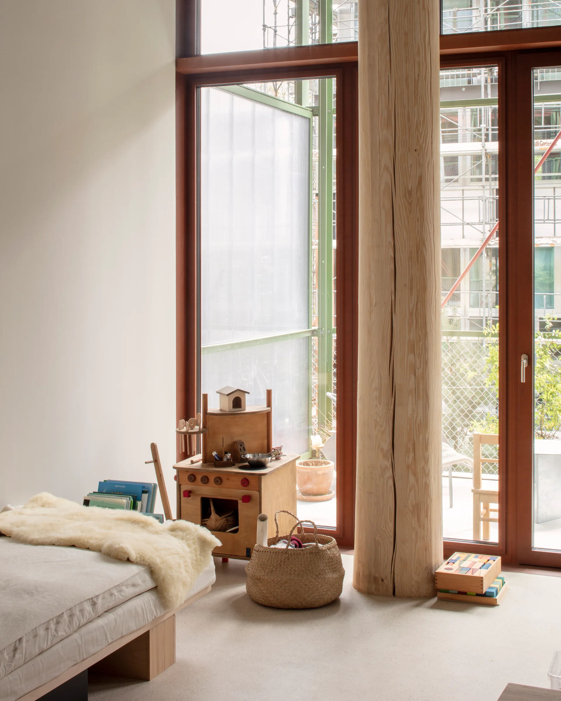 A child's bedroom with a low bed, a large natural wood column, a toy wooden kitchen, and glass doors leading to a balcony.