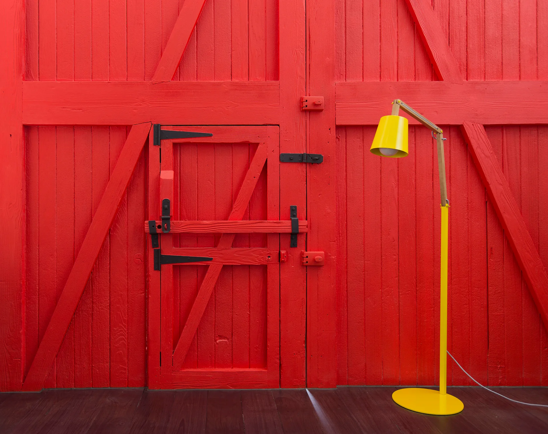 A bright yellow modern floor lamp stands on a dark wood floor in front of a rustic red painted barn door with black latches.