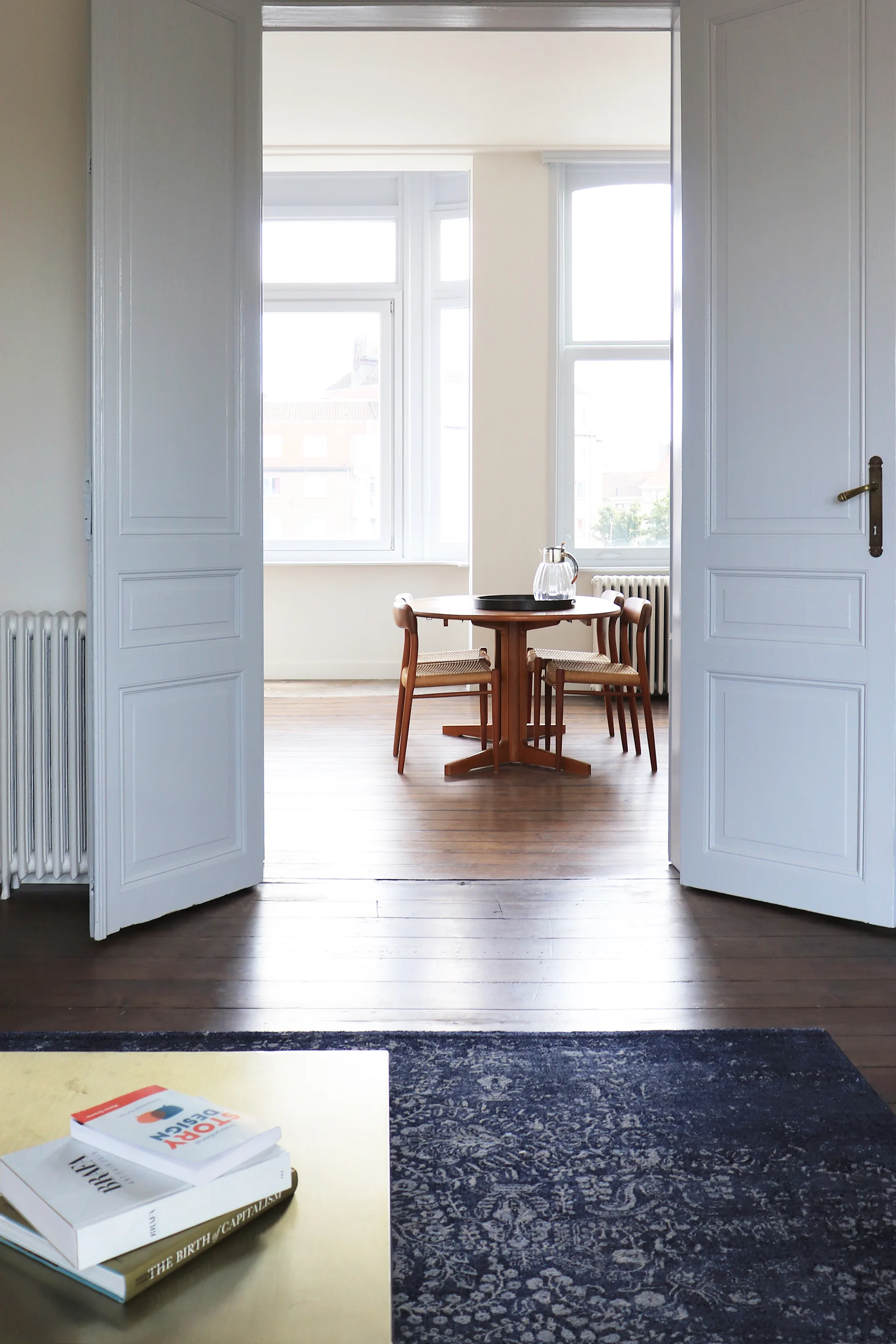 A view through open white double doors shows a mid-century modern wooden dining table and chairs set on hardwood floors in a sunlit room.
