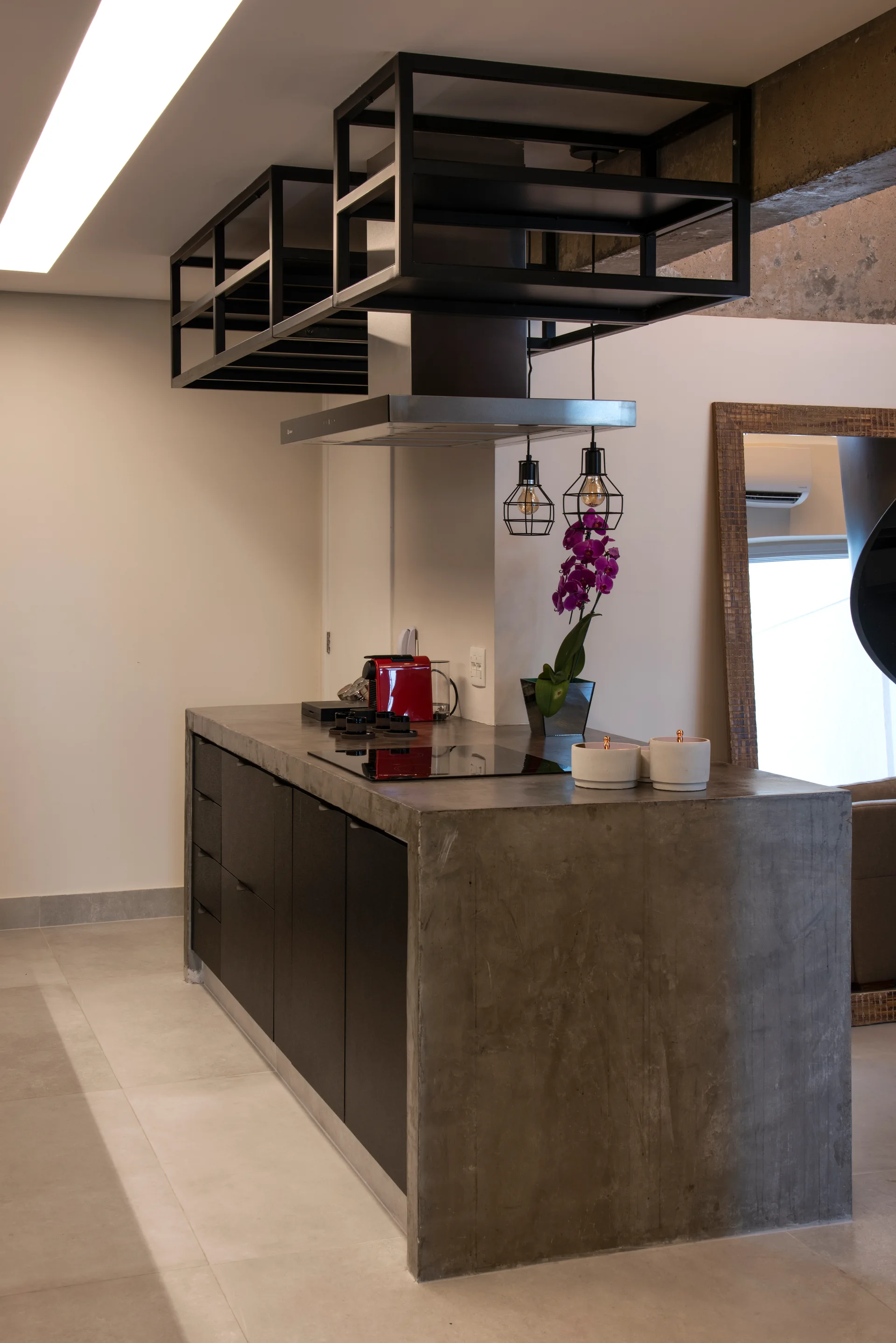 A concrete kitchen island with dark cabinetry sits below black metal ceiling-hung shelving and a metal range hood.