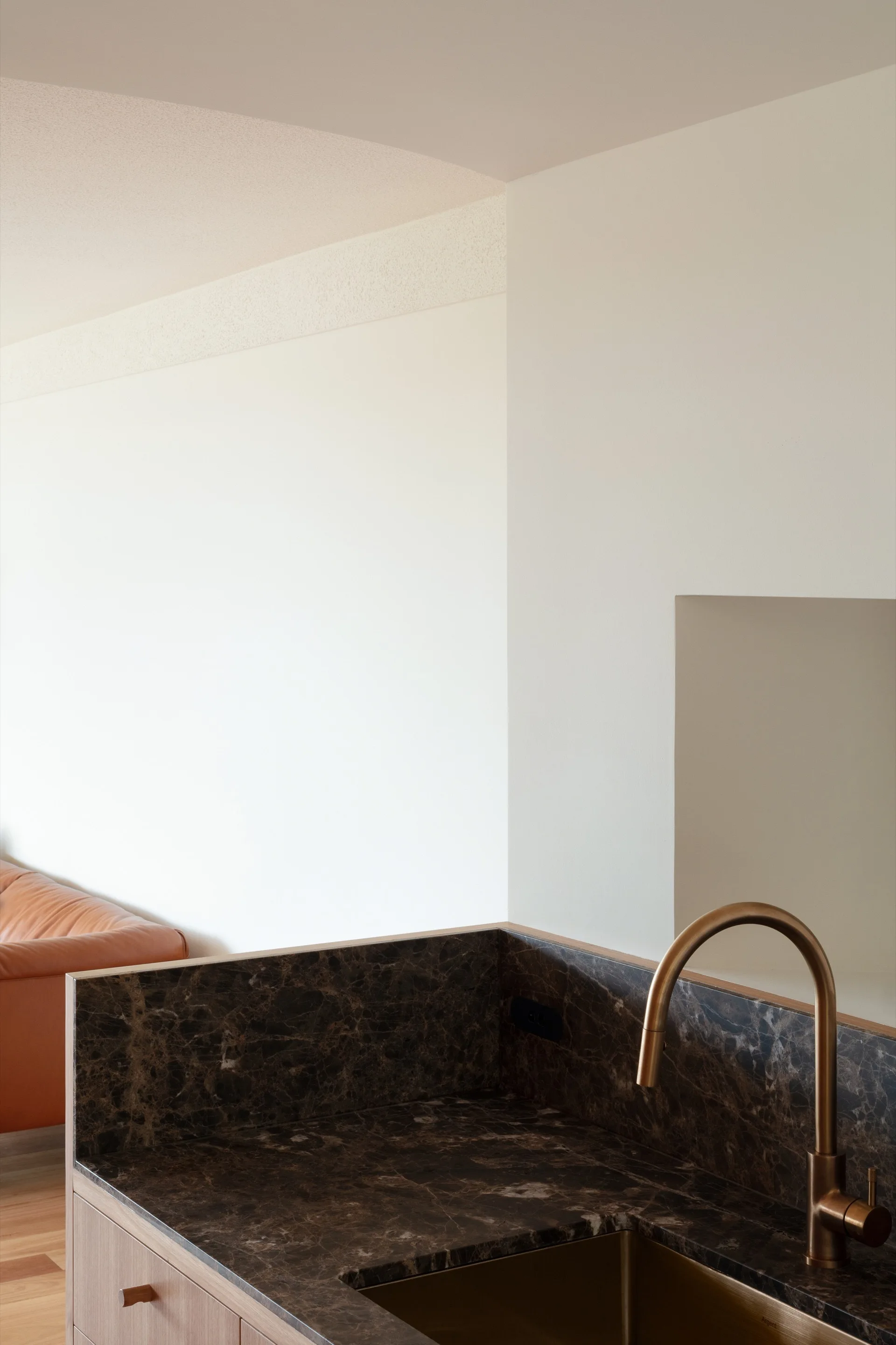 A close-up of a kitchen sink area featuring a dark brown marble countertop and brass faucet with a tan leather sofa visible in the background.