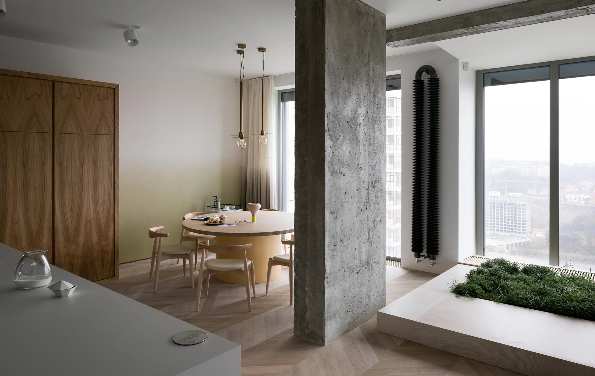 A minimalist dining room features a round wooden table, a central concrete pillar, herringbone floors, and a white-to-olive-green ombre wall.