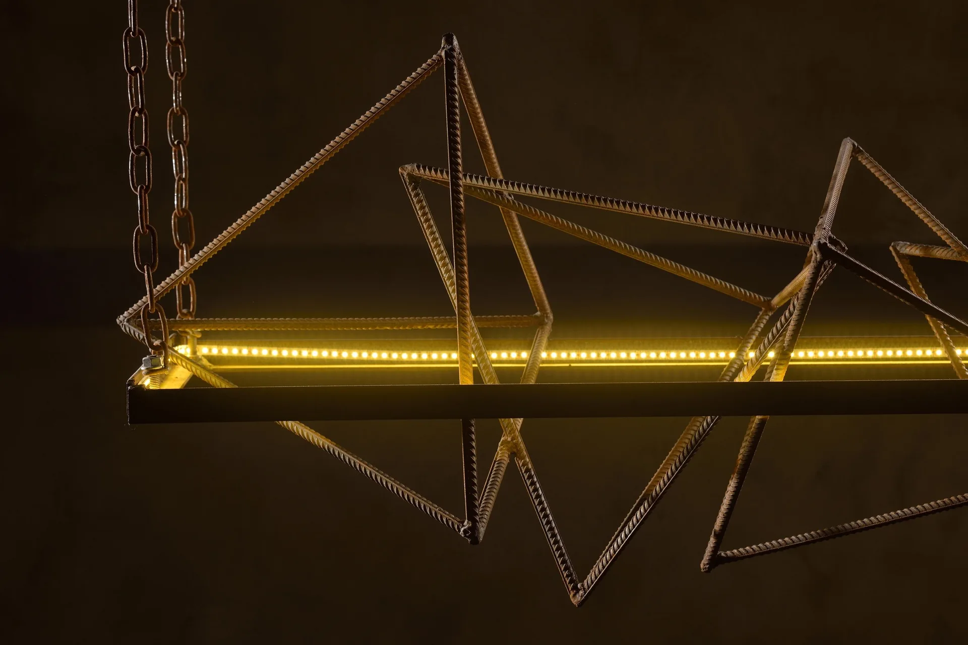 A sculptural industrial pendant light made of geometric rusted rebar with an integrated warm yellow LED strip suspended by a metal chain.